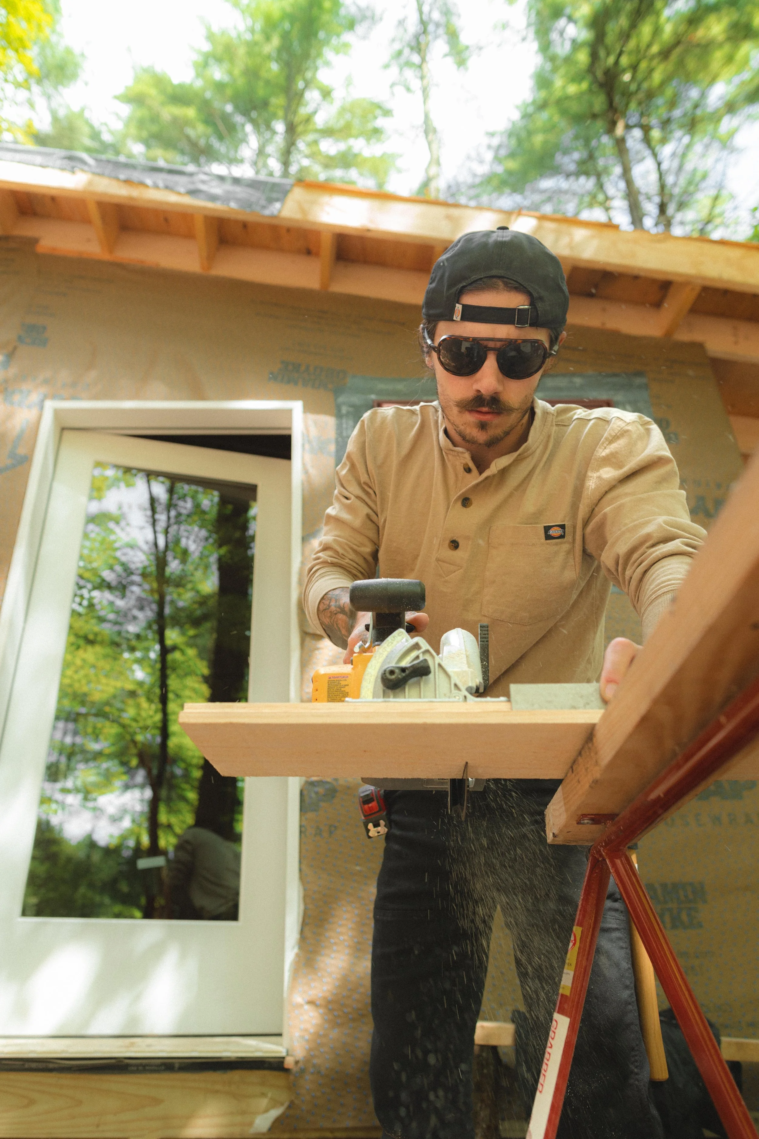 A man wearing sunglasses, a black hat backwards, and a beige shirt stands at a workbench using a power saw. The workbench has wood and tools, and he's focused on his work. There's a window and green trees outside, and part of a building under constru