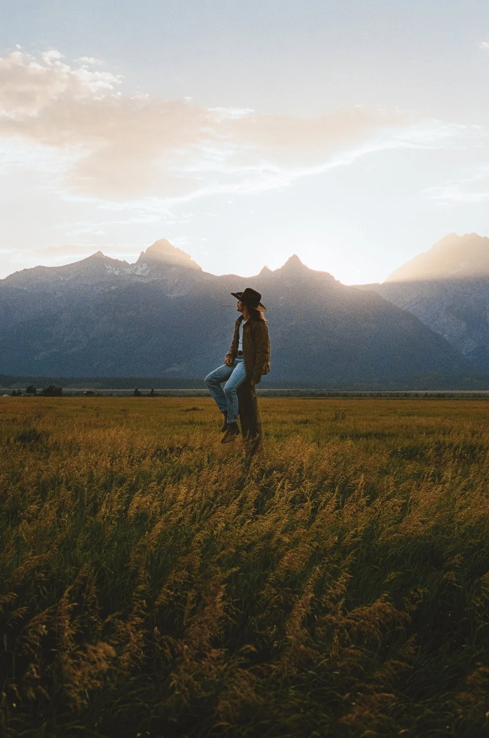 A woman in a black hat and jacket sits on a post in a field of tall grass with mountains and a cloudy sky in the background at sunset.