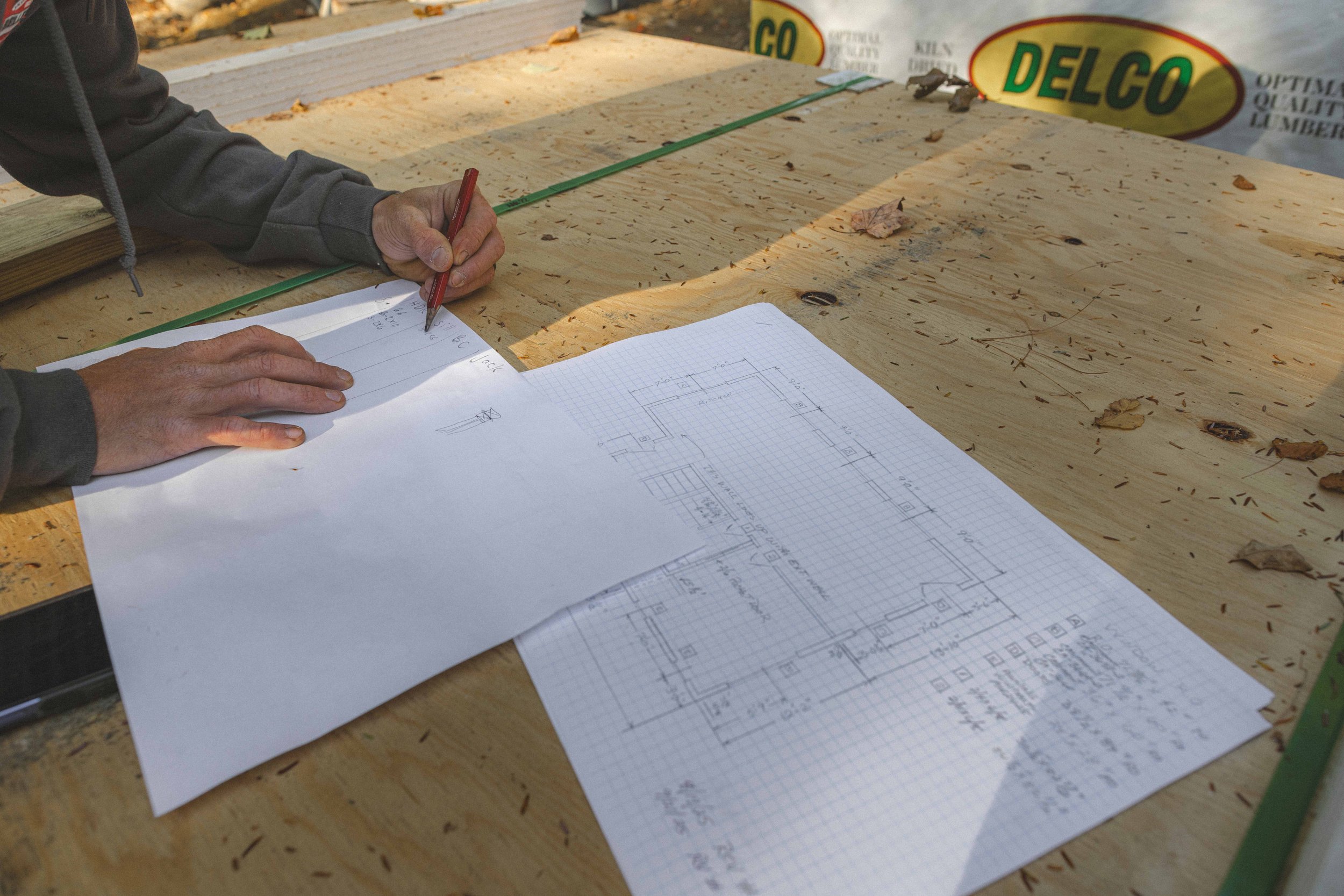 A person wearing a gray hoodie is drawing a floor plan on graph paper with a red pen, placed on a woodworking project table with scattered leaves and wood scraps. In the background, a banner displays the word 'DELCO'.