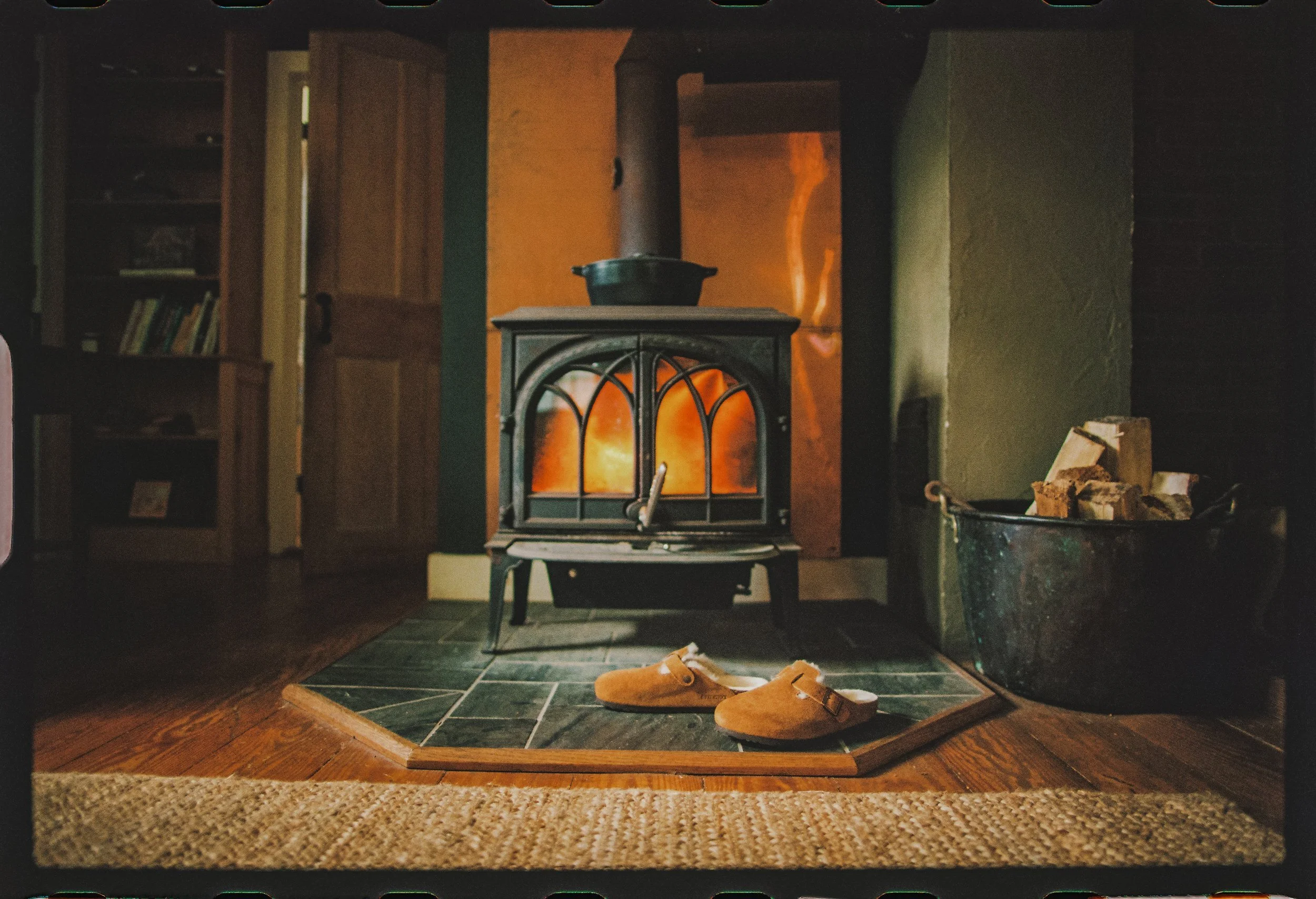 A cozy living room with a wood-burning stove in front of a tiled hearth, with a pair of tan slippers on the floor and a woven rug nearby. There's a stack of firewood in a metal bin to the right, a bookshelf with books to the left, and a glass door in the background with a warm glow.