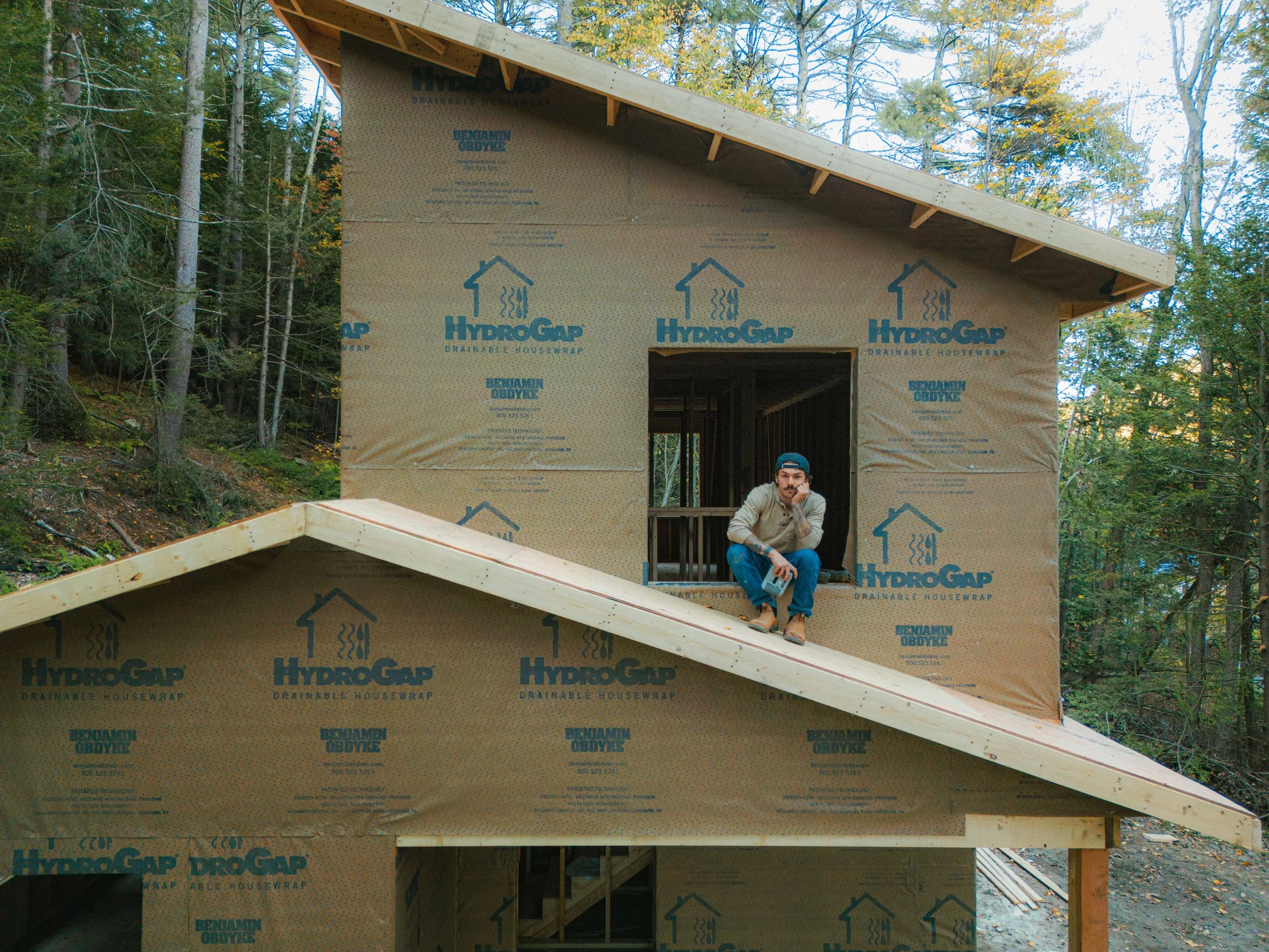 A man sitting on a wooden staircase in front of a house under construction during daytime. The house is wrapped in HydroGap house wrap and is set in a wooded area with tall trees.