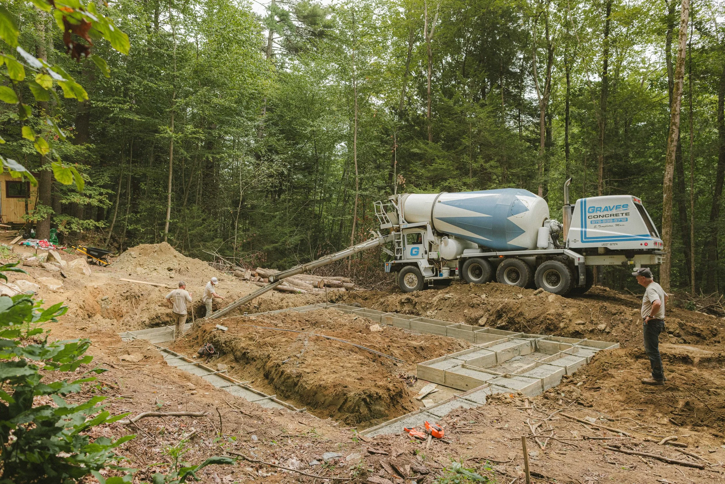 Construction site in a wooded area with a cement mixer truck pouring concrete into a foundation, workers guiding the process, and framing in progress.