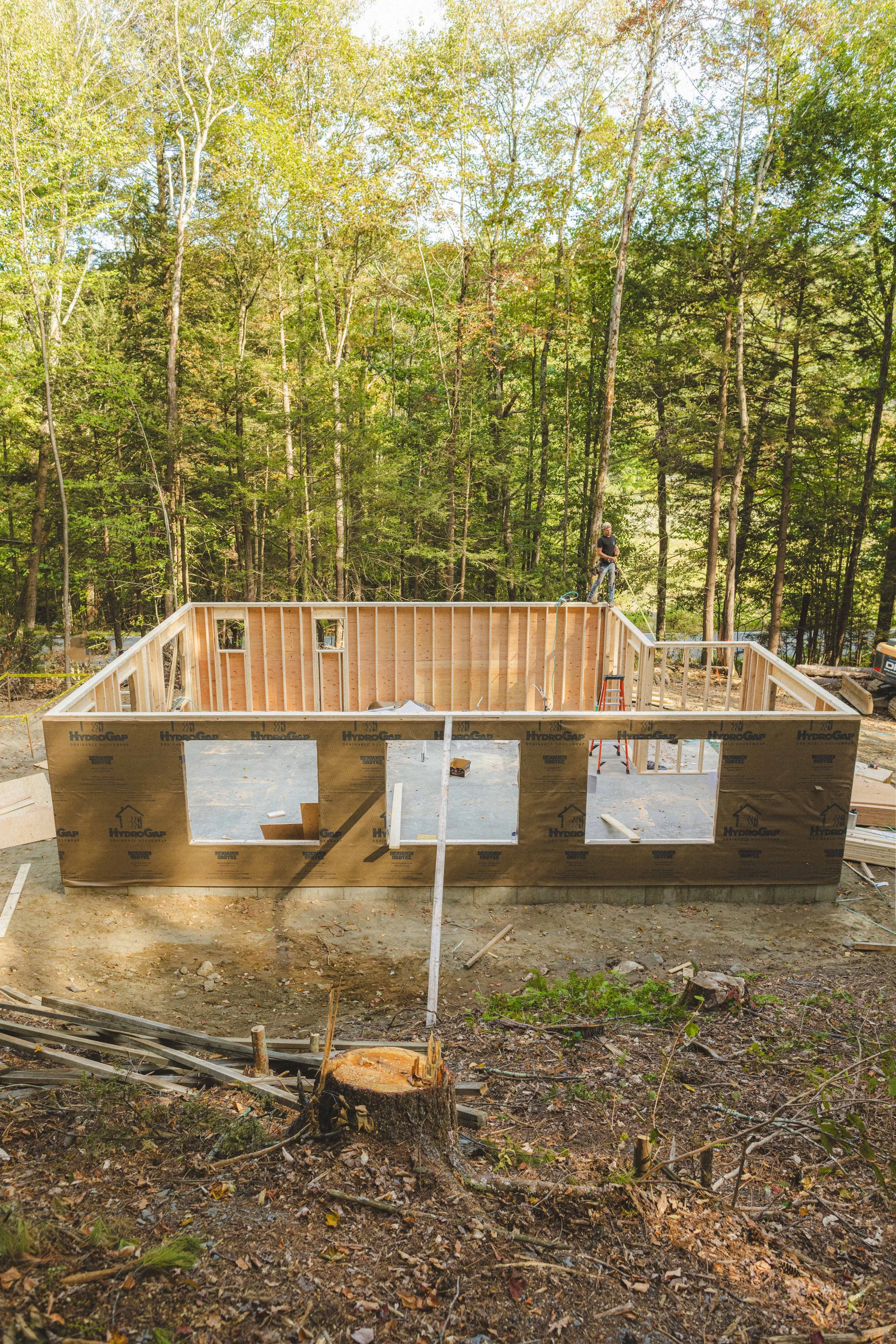 Construction site with a wooden framed house in progress surrounded by trees, with a man working on the roof.