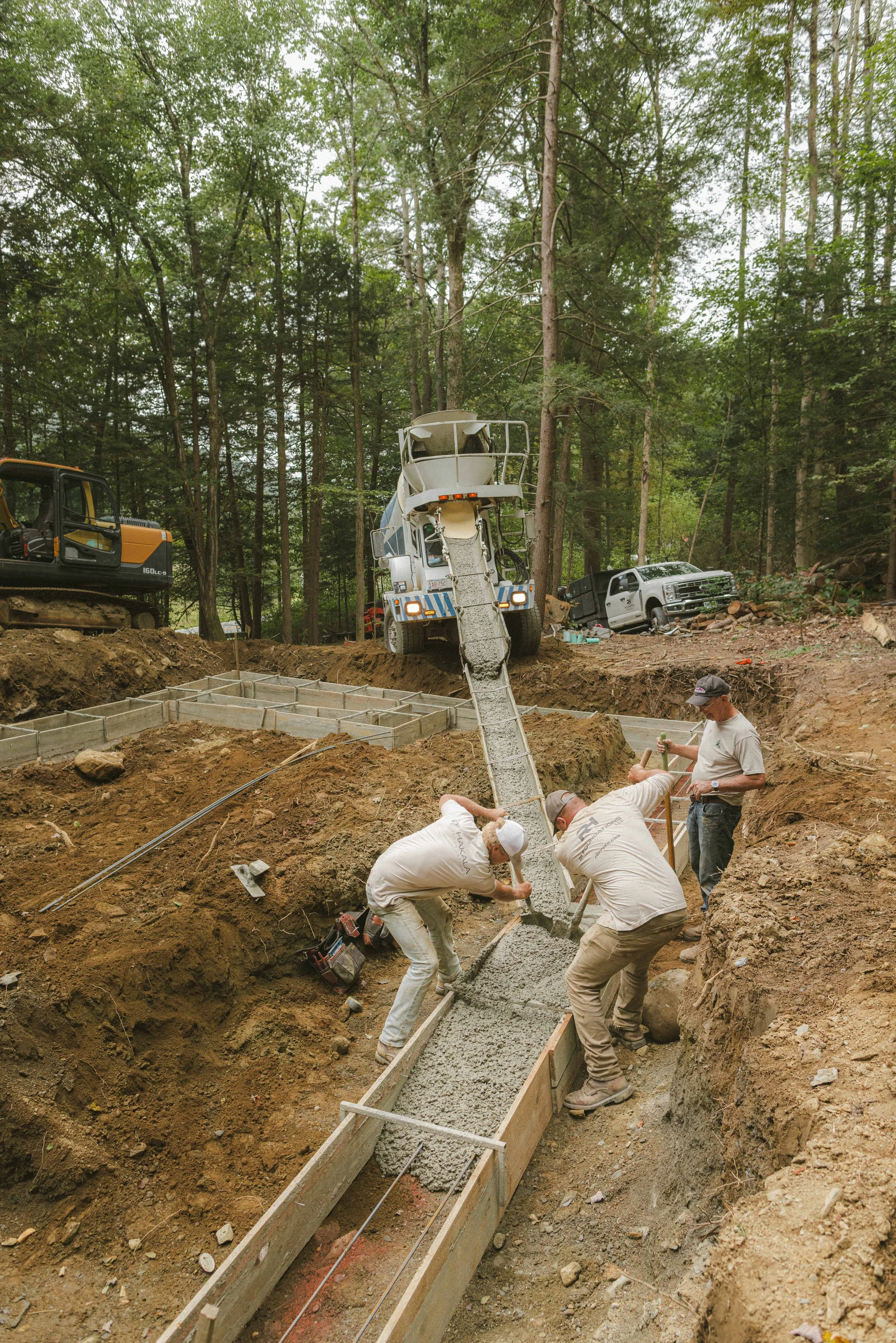 Workers pour concrete into a form for a foundation in a wooded area, with construction equipment and vehicles nearby.