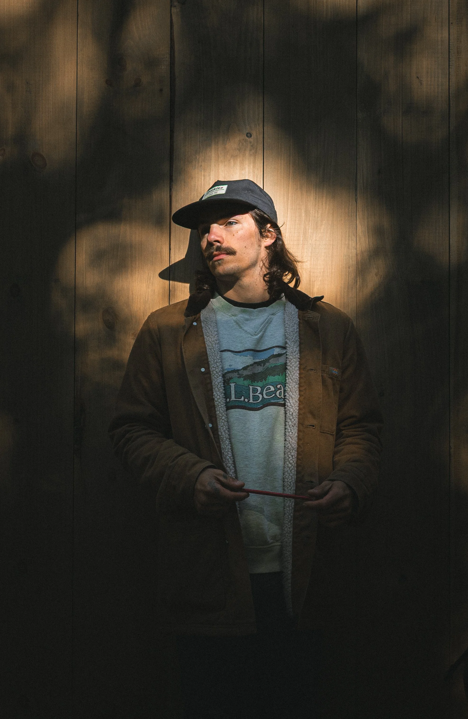 A man with long hair, mustache, wearing a black cap, brown jacket, and graphic t-shirt, standing against a wooden wall with diffused lighting creating shadows.