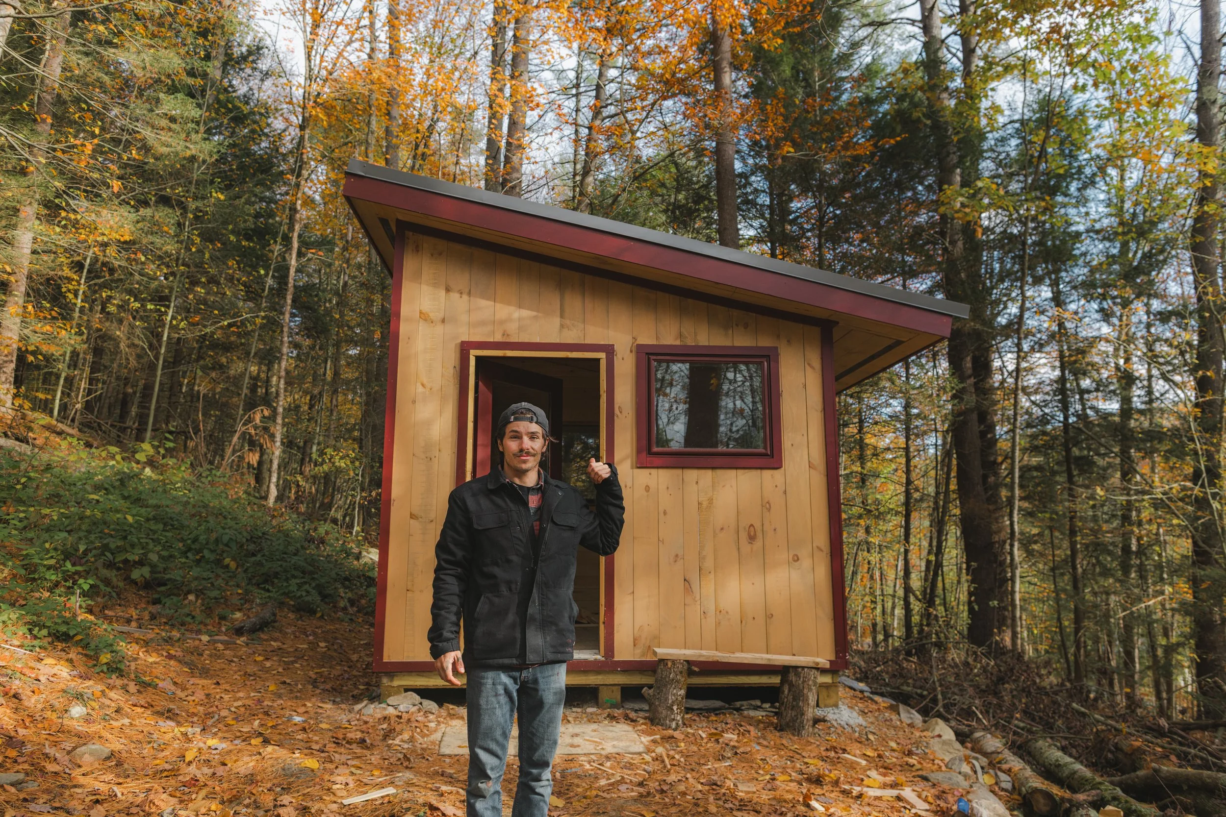 A man wearing a black jacket and gray jeans standing in front of a small wooden house in the woods during autumn.