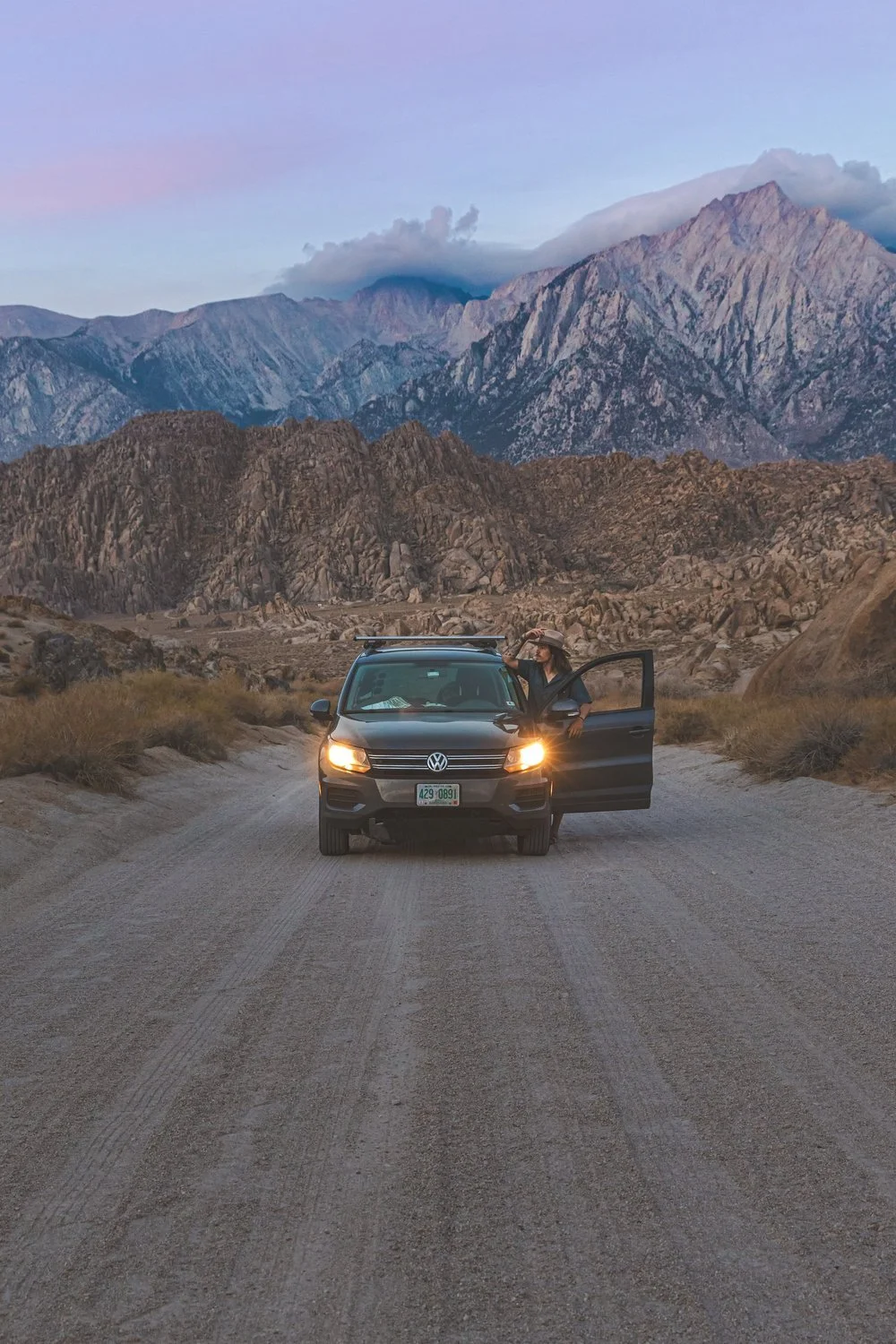 A woman standing beside a black Volkswagen SUV with its headlights on, parked on a dirt road amidst a mountainous landscape during twilight, with mountains and clouds in the background.