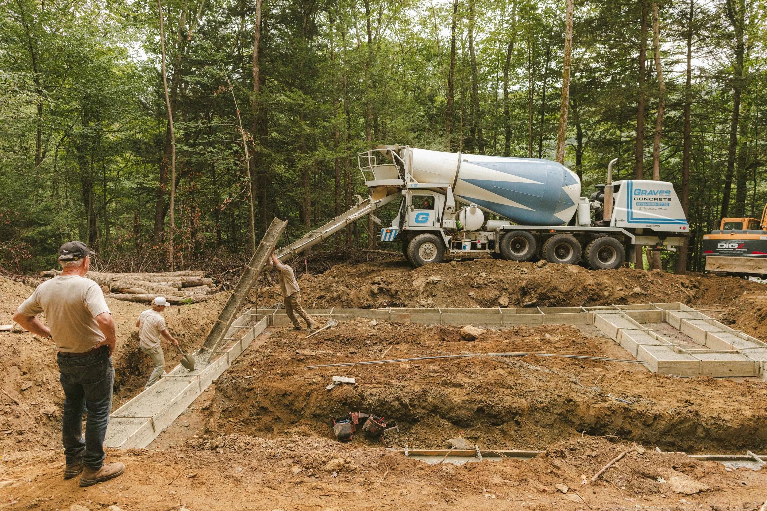 Construction site in a wooded area with workers and a cement mixer truck preparing foundations for a building.