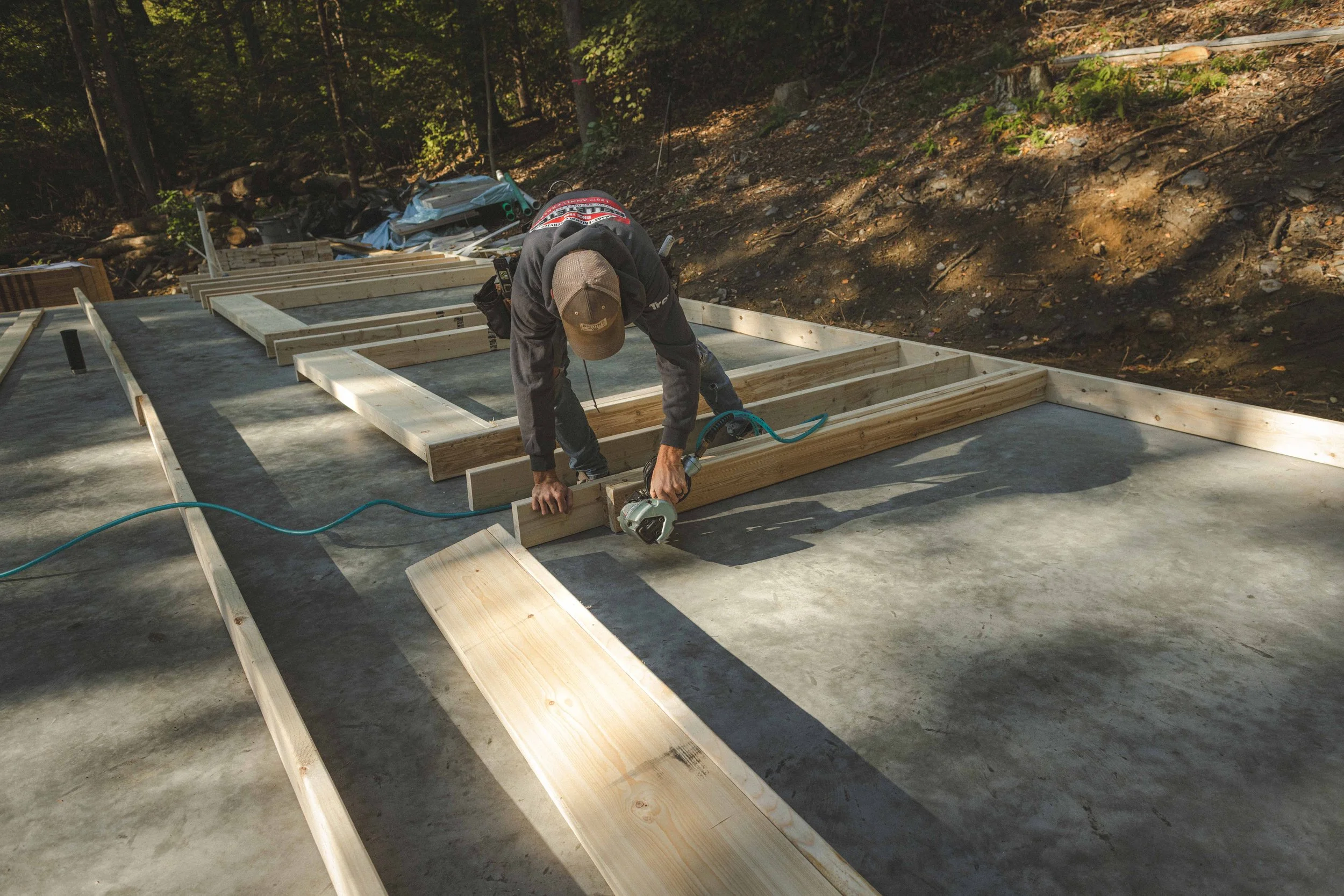 A construction worker installing wooden framing on a concrete foundation outdoors during daytime, surrounded by trees and construction debris.