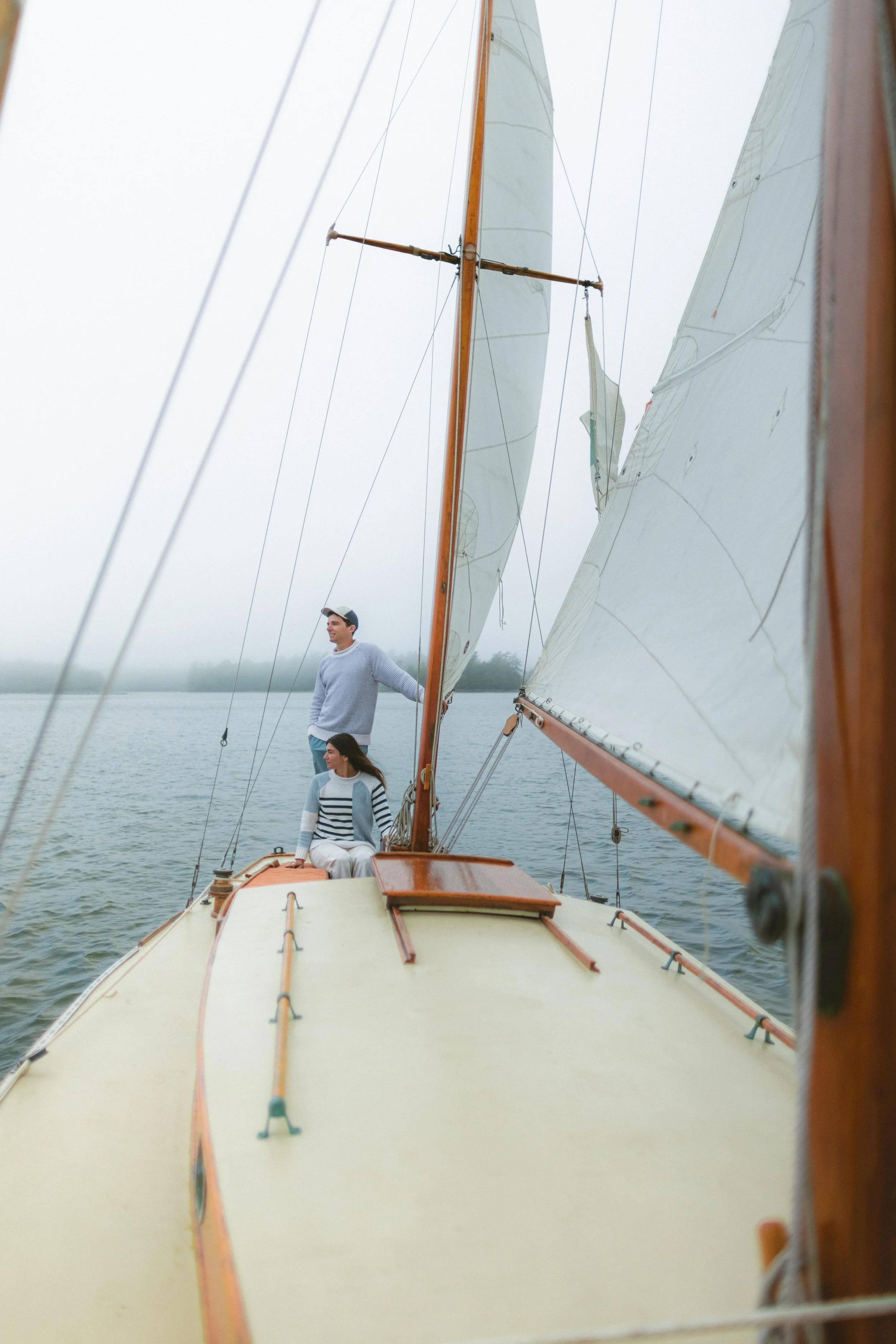 People sailing on a boat in a foggy body of water, with visible sails and wooden mast, two women seated on the boat's deck and a man standing.