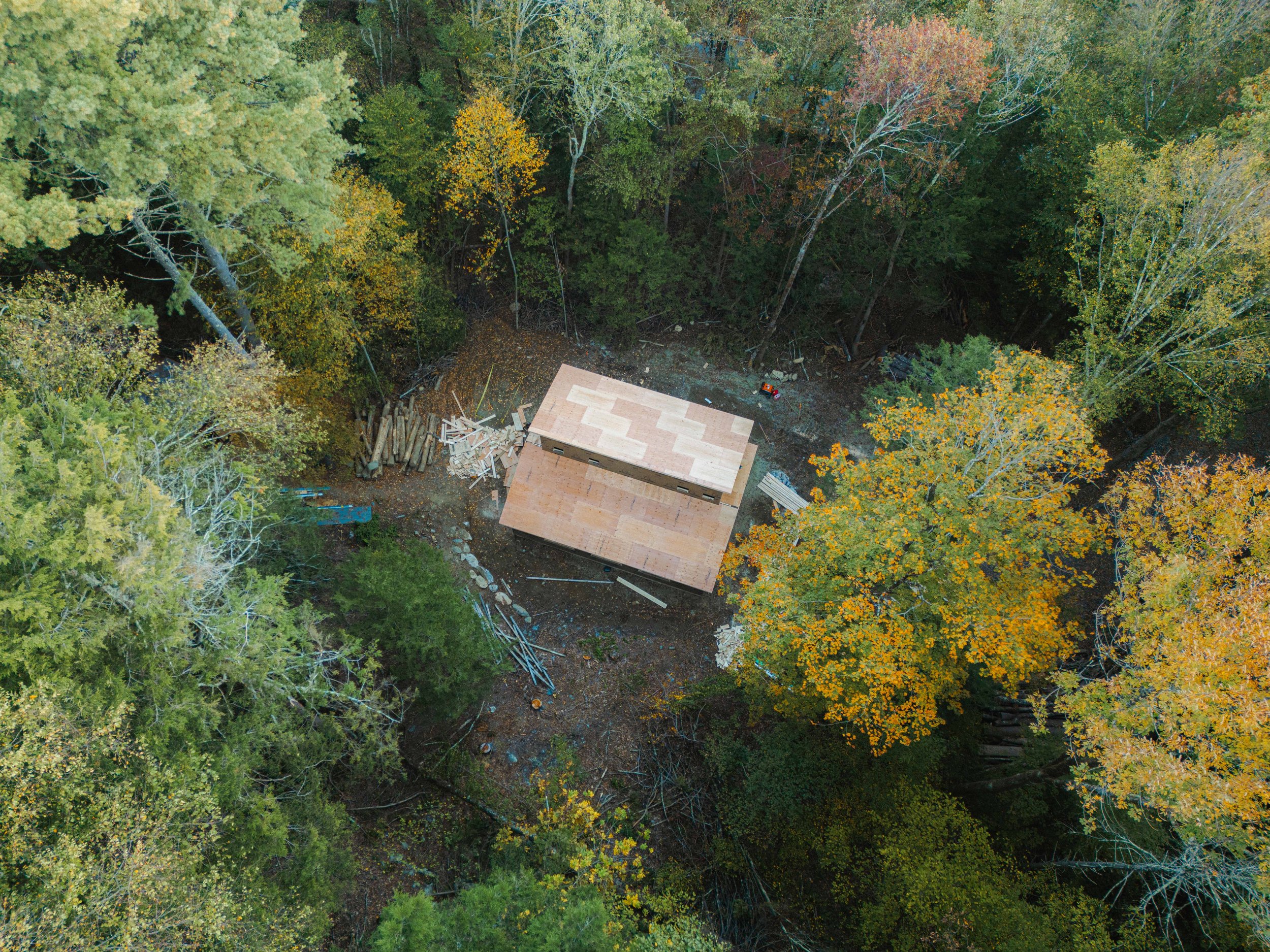 Aerial view of a construction site in a forest with trees displaying fall colors, a partially built wooden structure, construction materials, and equipment.