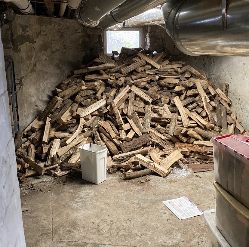 A room with a large pile of chopped firewood stacked against the back wall, with a small window above it. There is a white container with a handle in front of the wood pile, and what appears to be storage boxes on the right side. Metal pipes run along the ceiling, and the floor is concrete.