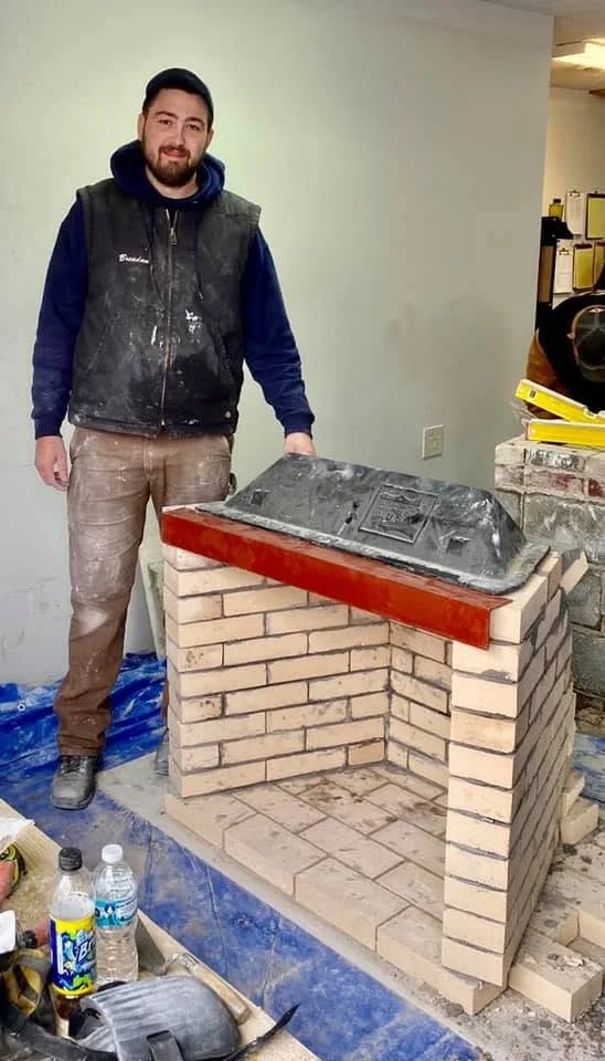 A man standing next to a masonry fireplace under construction, inside a room with construction materials and tools around. Work done by Two River Chimney Services serving Monmouth and Ocean County, NJ
