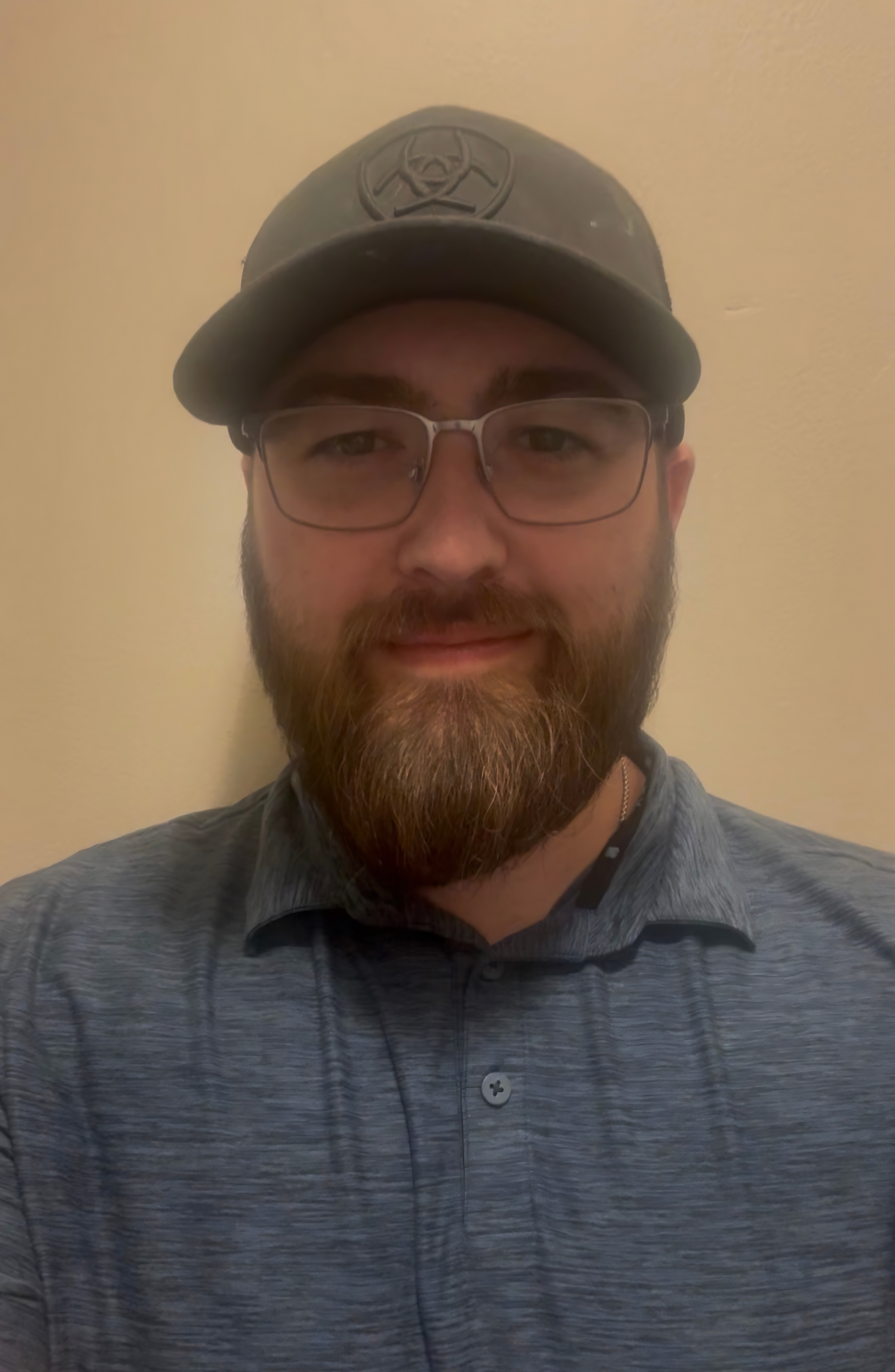 A man with glasses and a beard wearing a button-up shirt and a baseball cap with a logo, smiling and looking at the camera, against a plain light-colored background. Owner of Two River Chimney Services serving Monmouth and Ocean County, NJ