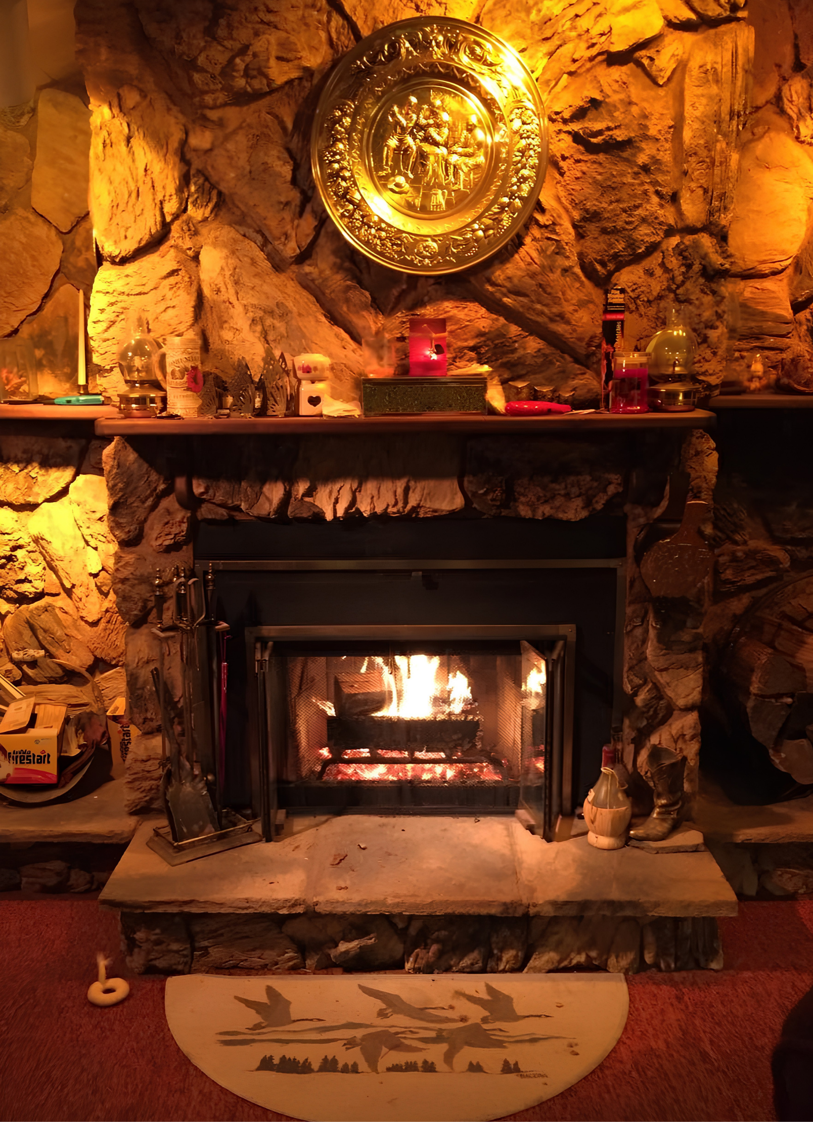 A cozy stone fireplace with a fire burning inside, decorated with various candles, ornaments, and small decorative objects on the mantel. A wooden picture with silhouette of flying birds is placed in front of the fireplace.