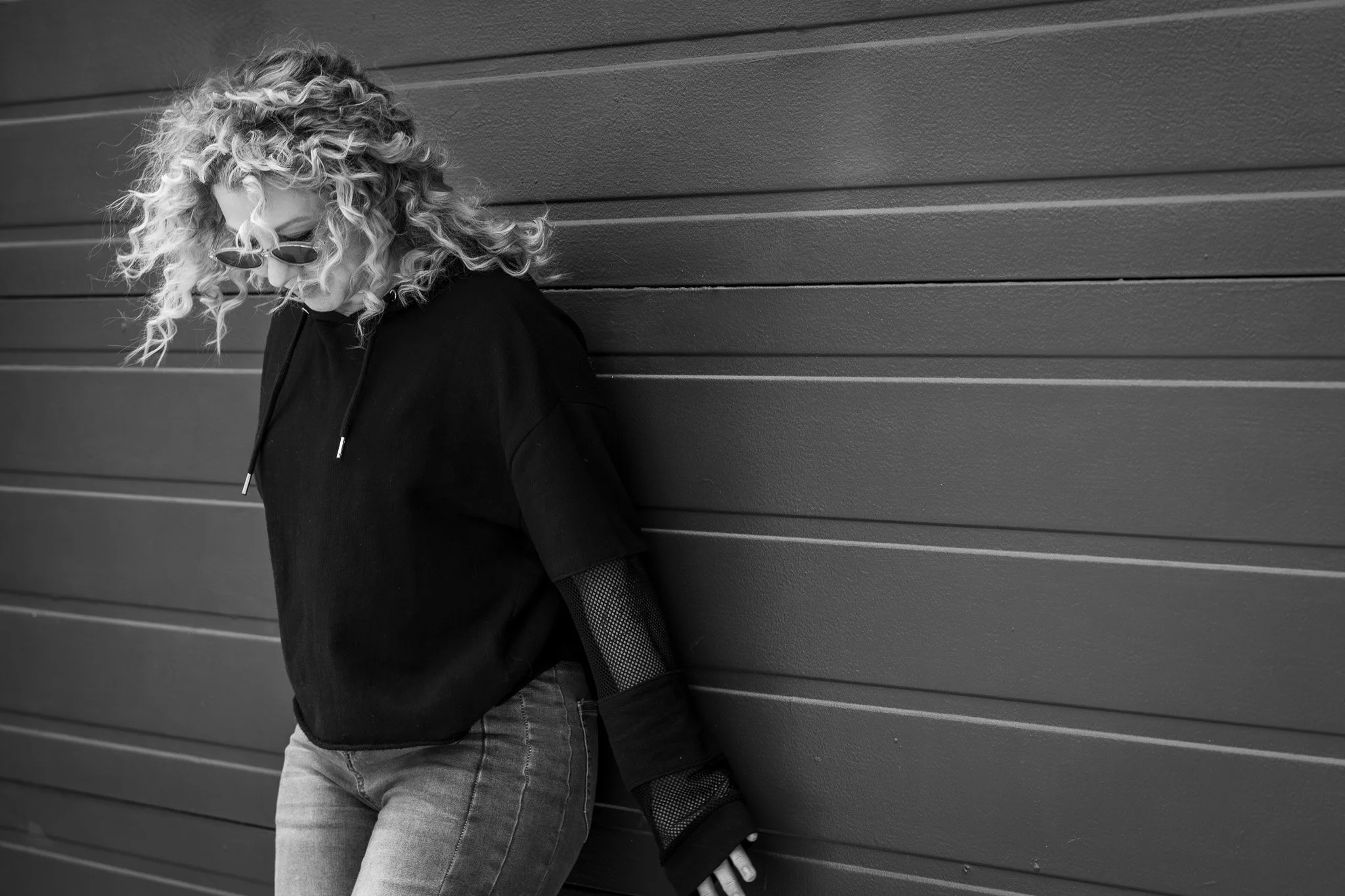 Black and white photo of a woman with curly hair wearing round sunglasses and a black hoodie, leaning against a wooden wall.