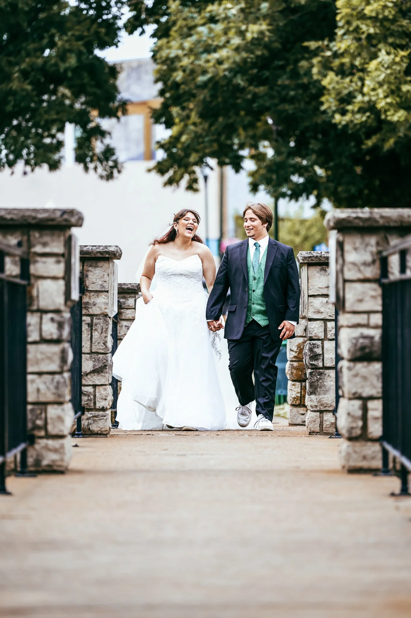 A bride and groom walking hand in hand through a stone archway, smiling and laughing outdoors during their wedding day.