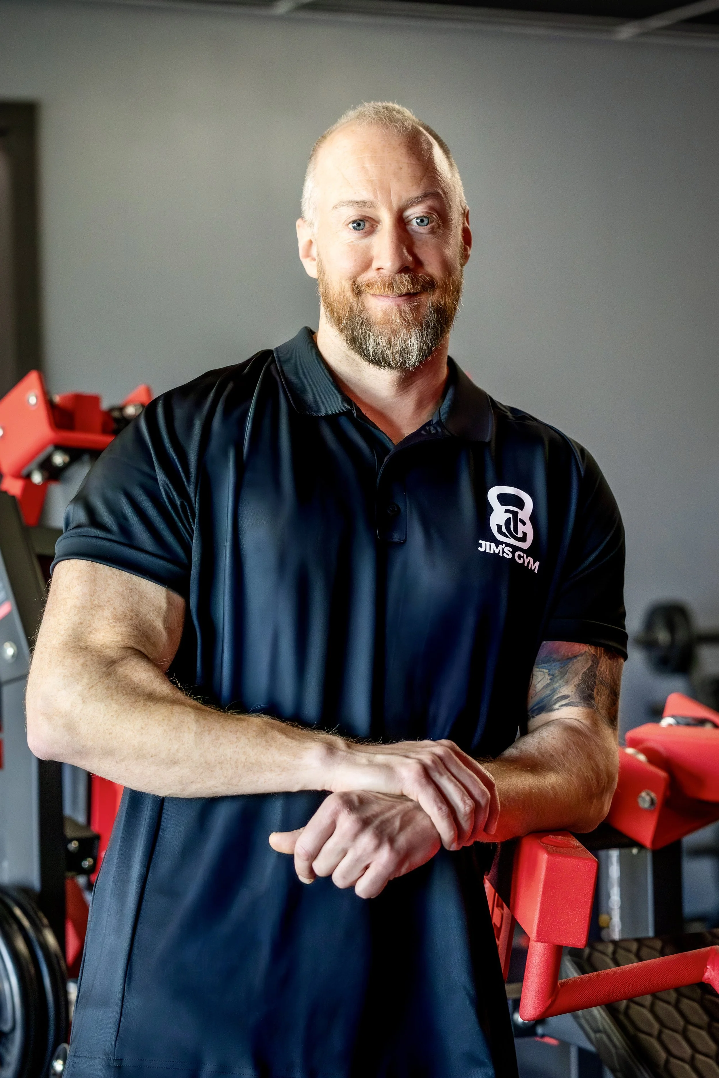 A man with a beard and blue eyes, wearing a black polo shirt with a logo that says 'Jim's Gym,' standing in a gym with red and black equipment in the background.