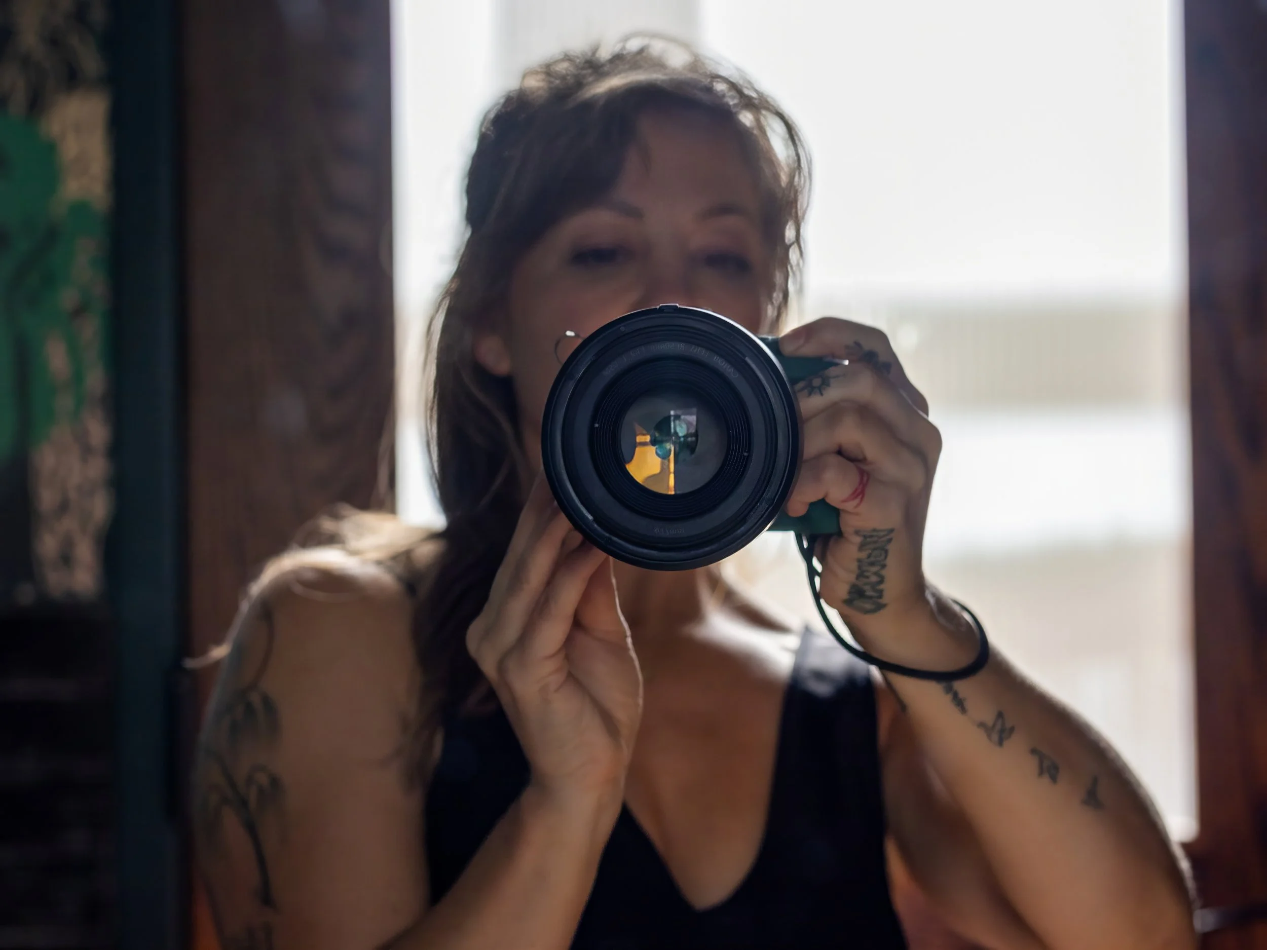 A young woman with curly hair is taking a mirror selfie using a camera, showing her face and tattoos on her arms.