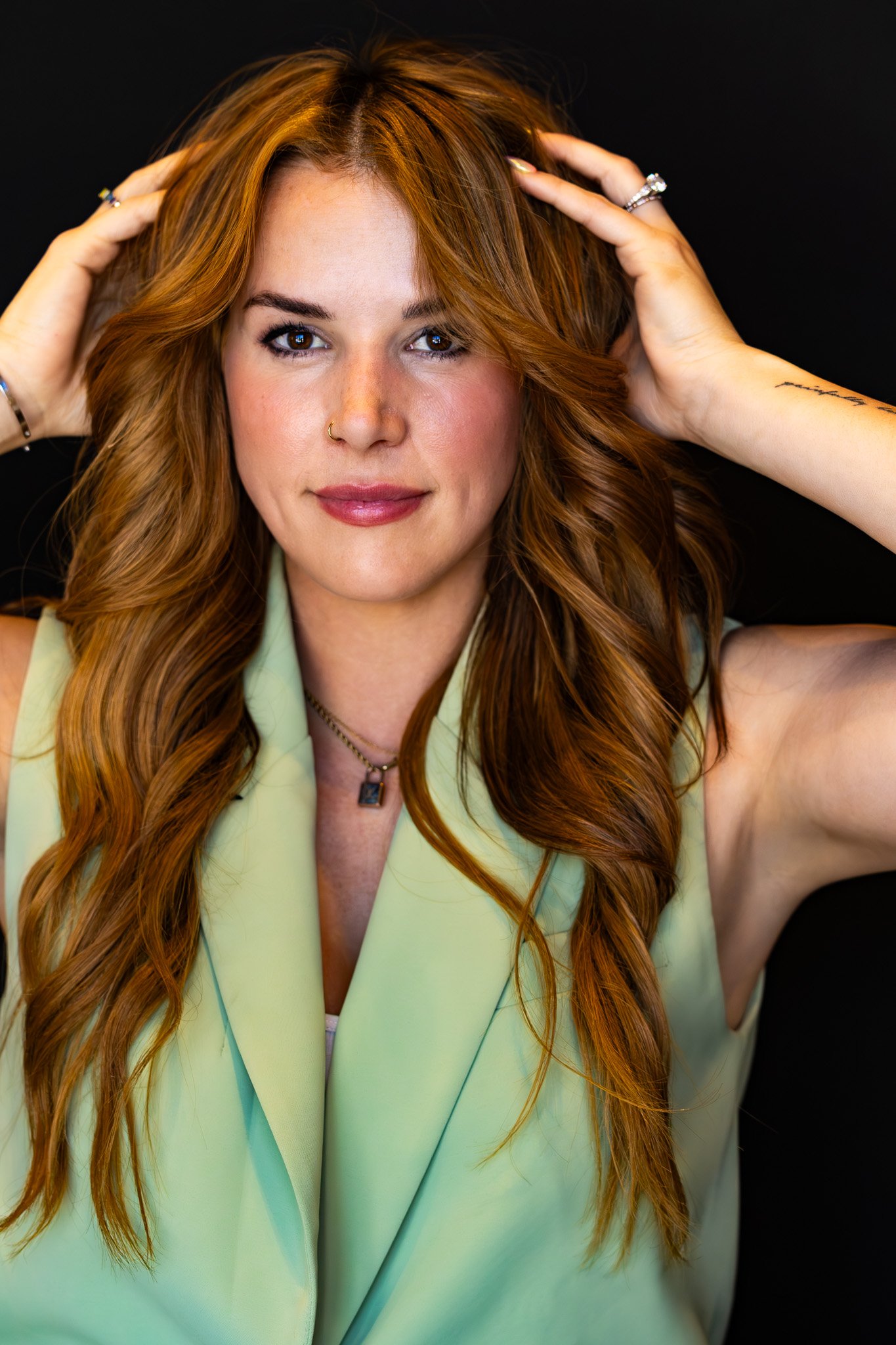 A woman with long, curly, red hair holding her hands on her head, wearing a light green sleeveless blazer, jewelry including rings and necklaces, and a nose ring, against a black background.