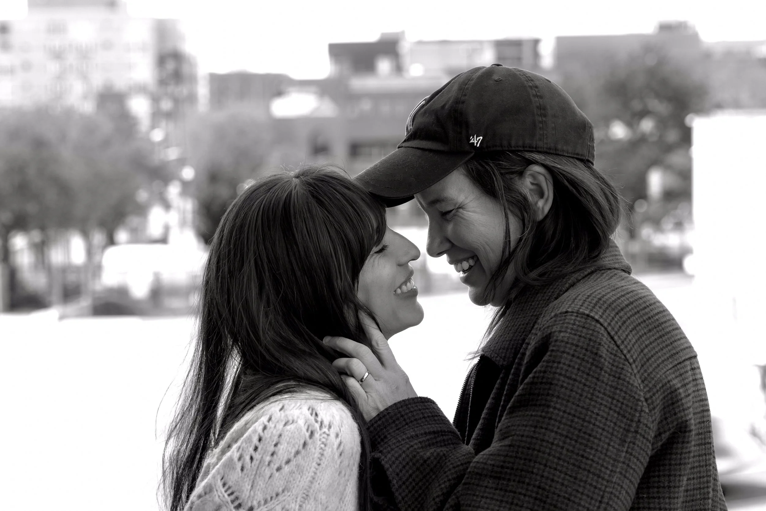 A black-and-white photo of a smiling woman with a ponytail and wearing a knit sweater, and a woman in a baseball cap and jacket, gently touching foreheads and smiling outdoors with blurred buildings and trees in the background.