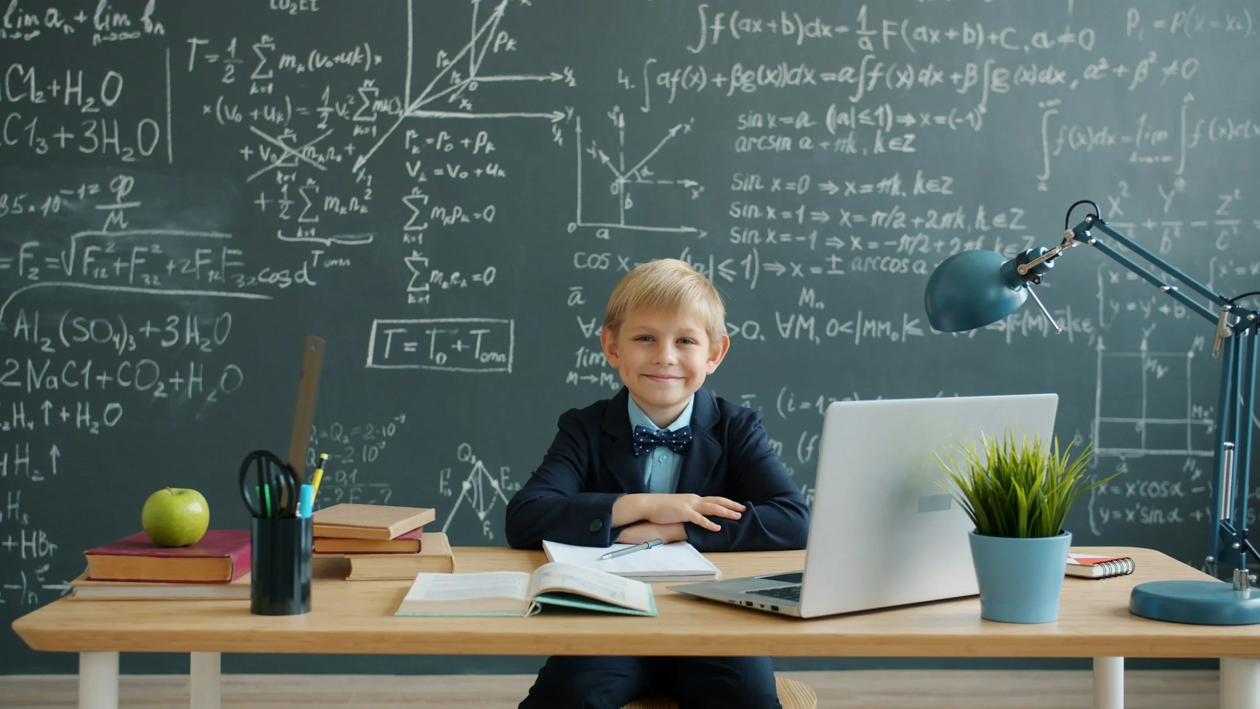 A young boy dressed in a suit and bow tie sits at a wooden desk with arms crossed, smiling at the camera in front of a chalkboard filled with math and science formulas. The desk has a green apple, books, a potted plant, and stationery, with a desk lamp, laptop, and open workbook.