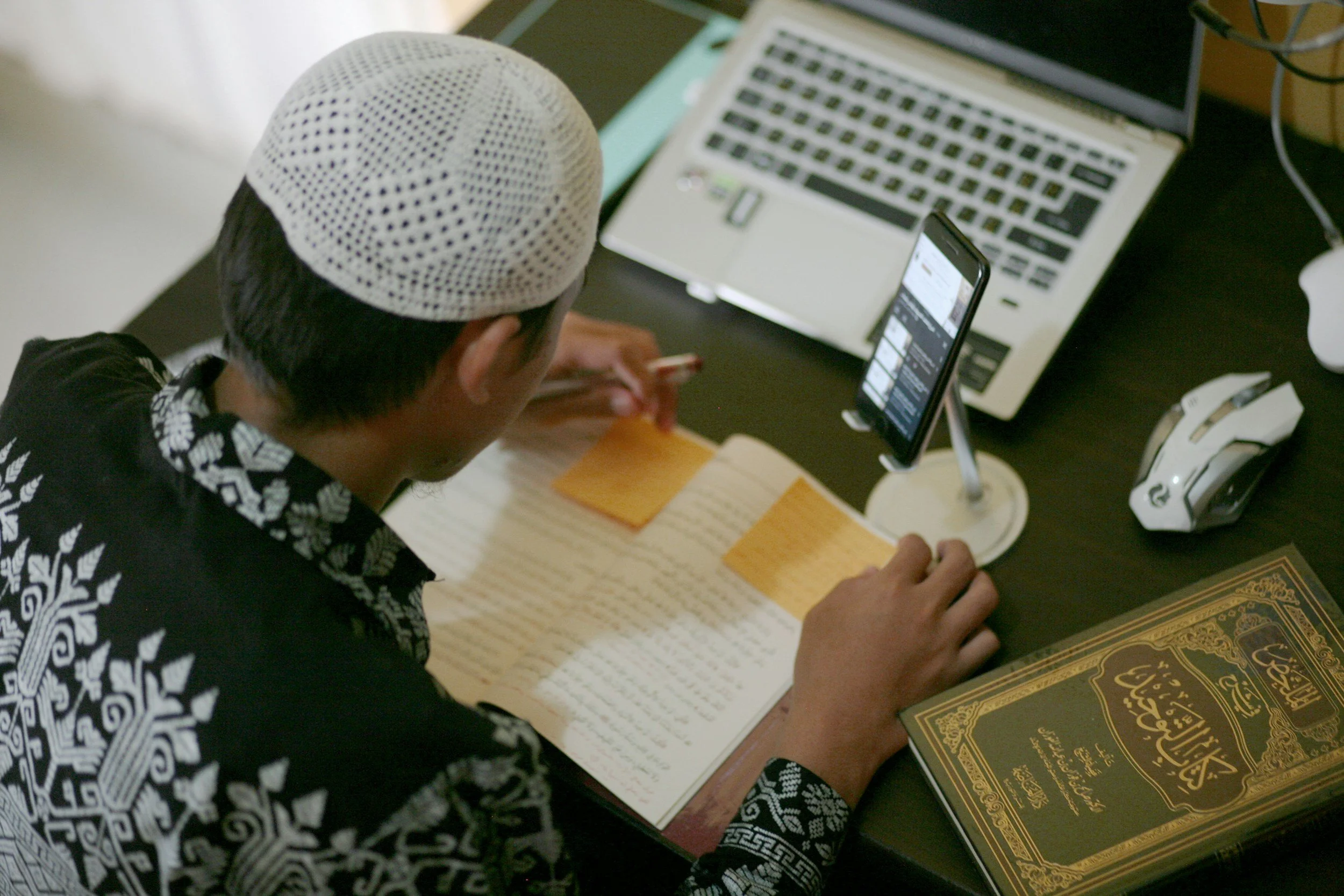 A person wearing a white knit cap and a black and white patterned shirt, reading a book with Arabic text, with a laptop, a smartphone, and other items on the desk.