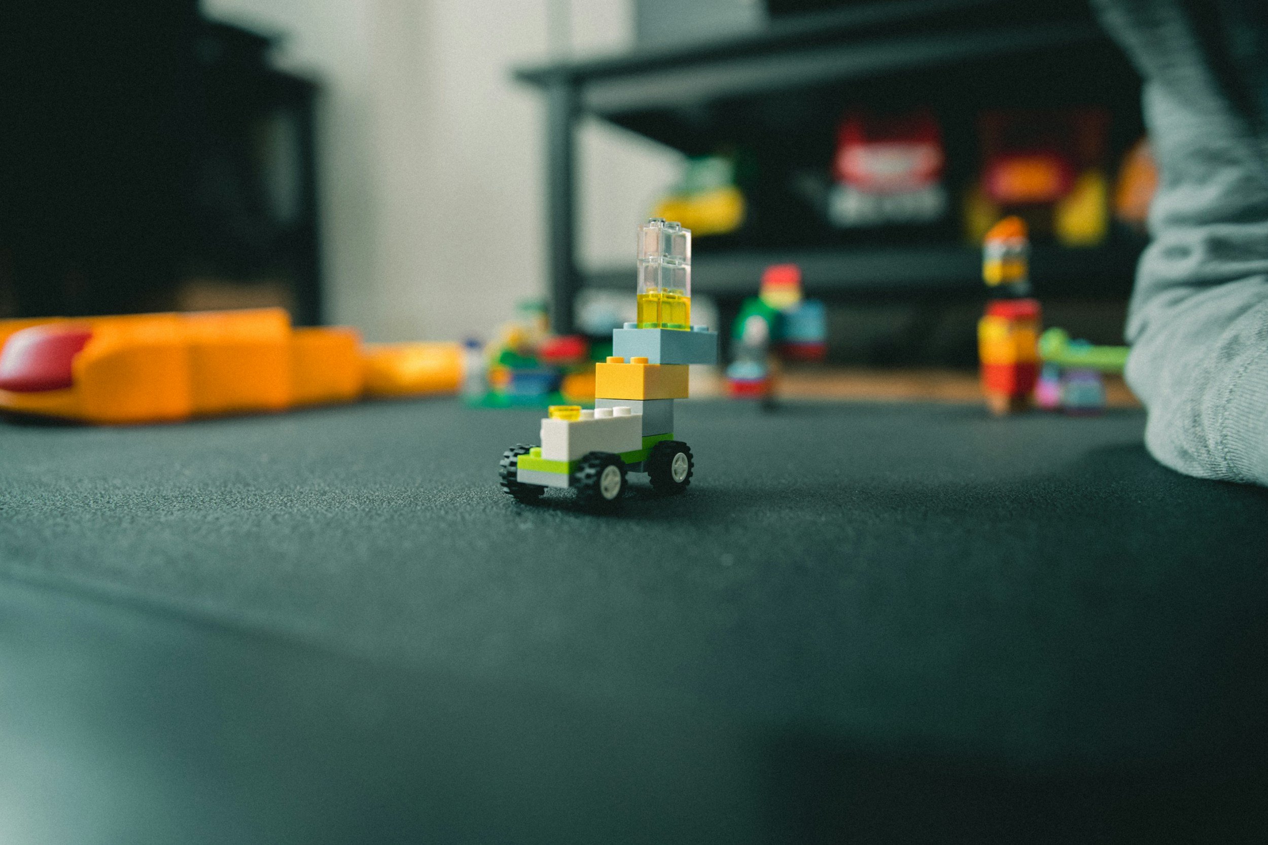 A small LEGO vehicle with white, yellow, and green bricks and a transparent yellow piece on top, on a dark surface with blurred colorful LEGO pieces and toys in the background.