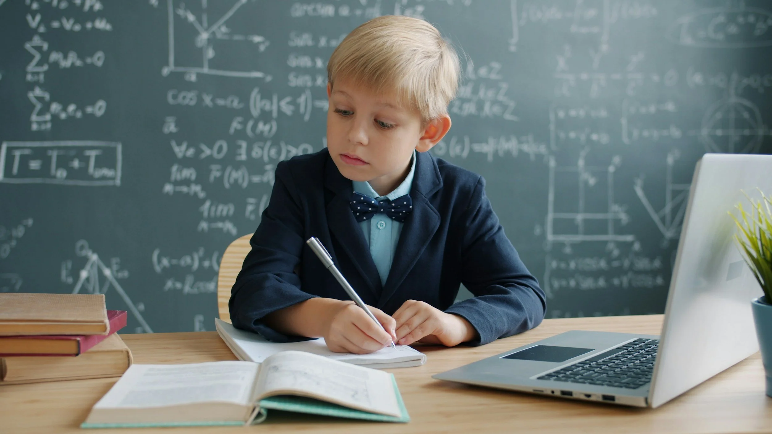 Young boy in a navy blazer writing in a notebook at a wooden desk surrounded by books, with scientific formulas on a chalkboard behind him and an open laptop to his side.