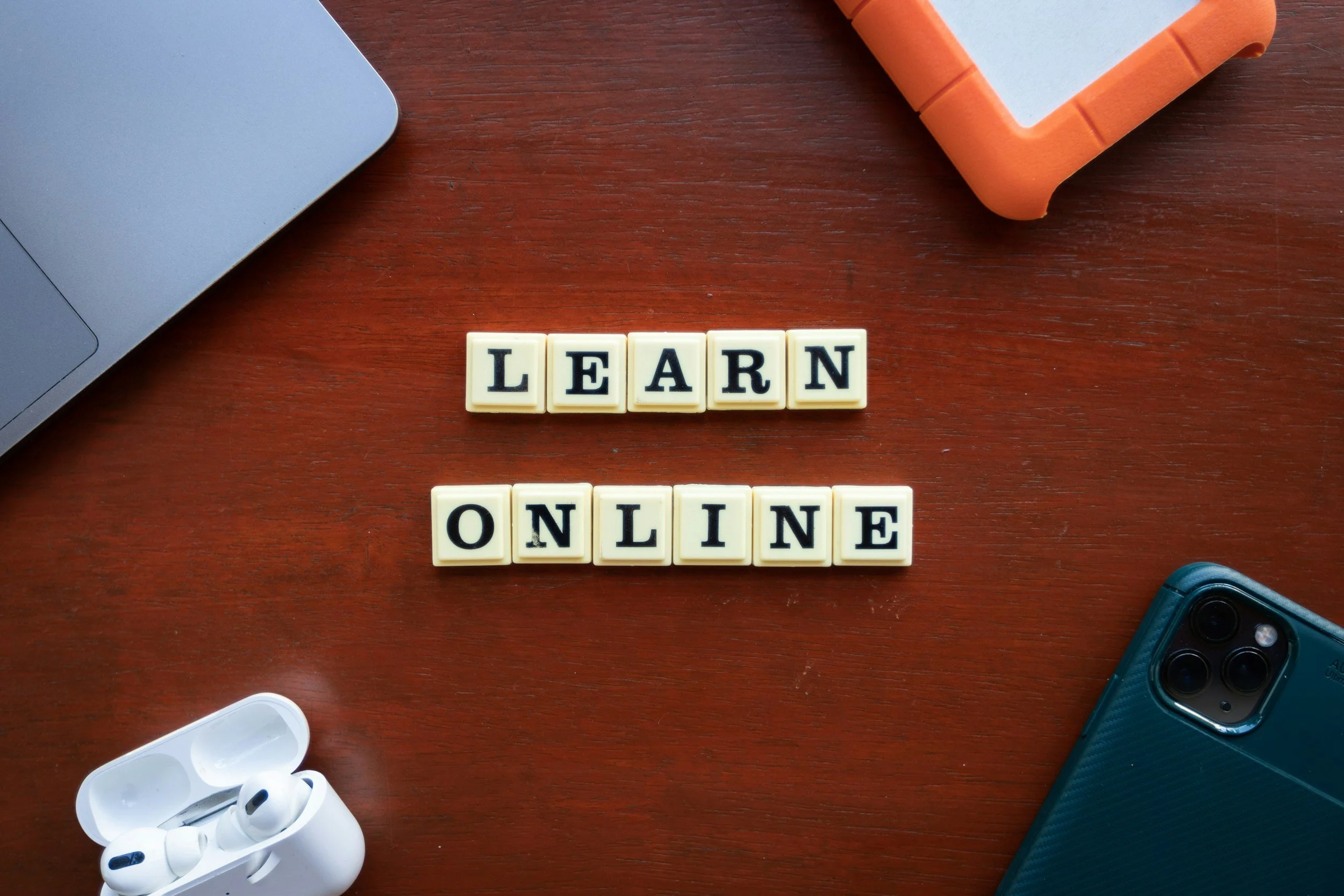 Wooden desk with white letter tiles spelling out "LEARN ONLINE", wireless earbuds in an open case, part of a laptop, a sports camera, and a foam microphone windscreen.