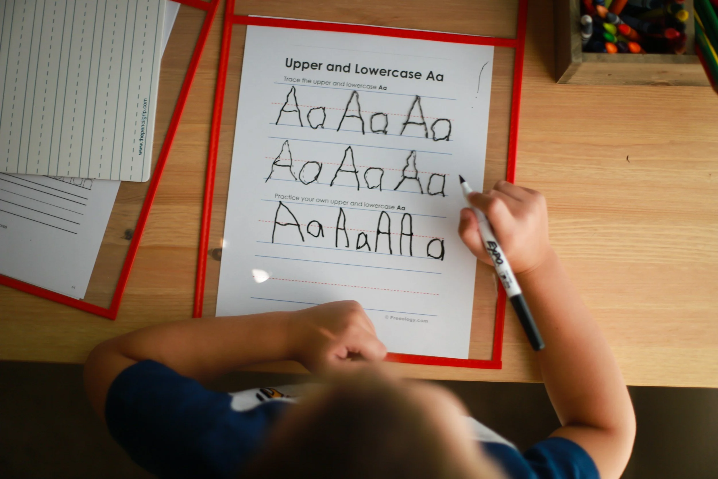 Child practicing writing uppercase and lowercase letter A on a worksheet, using a black marker, on a wooden desk surrounded by school supplies.