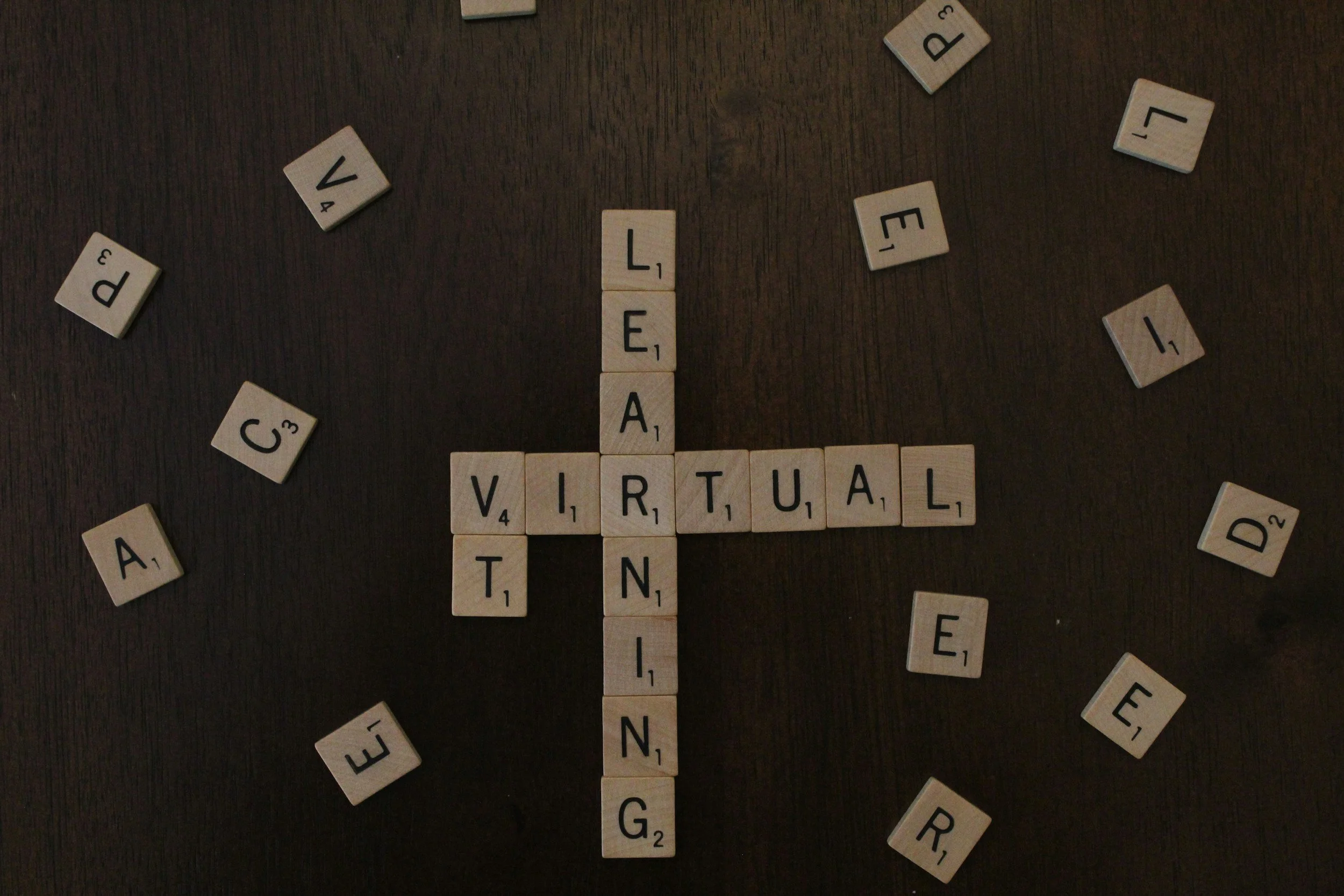 Scrabble tiles spelling 'VIRTUAL' and forming a cross with tiles spelling 'PEACE' and other scattered letter tiles on a dark wooden surface.