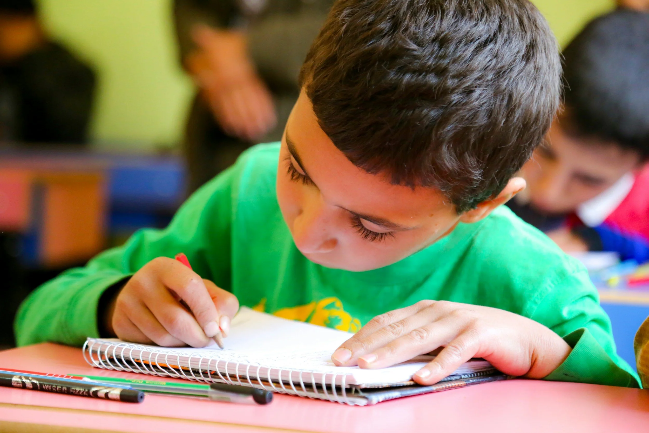 A young boy with dark hair, wearing a bright green shirt, is focused on writing or drawing on a spiral notebook at a pink table in a classroom, with another child blurred in the background.
