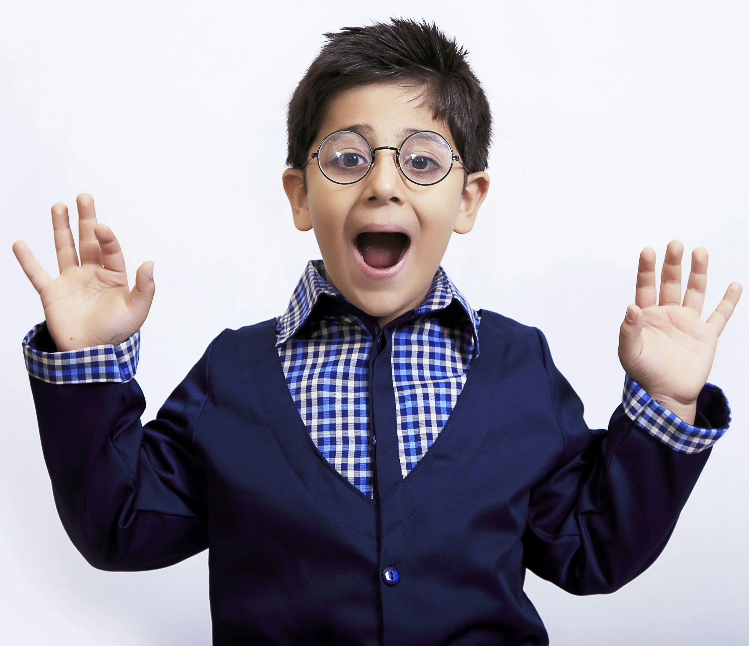 A young boy with short, dark hair, wearing round glasses, a blue shirt with a checkered collar, and a dark blazer, has his mouth open and hands raised with fingers spread. He appears surprised or excited against a plain white background.