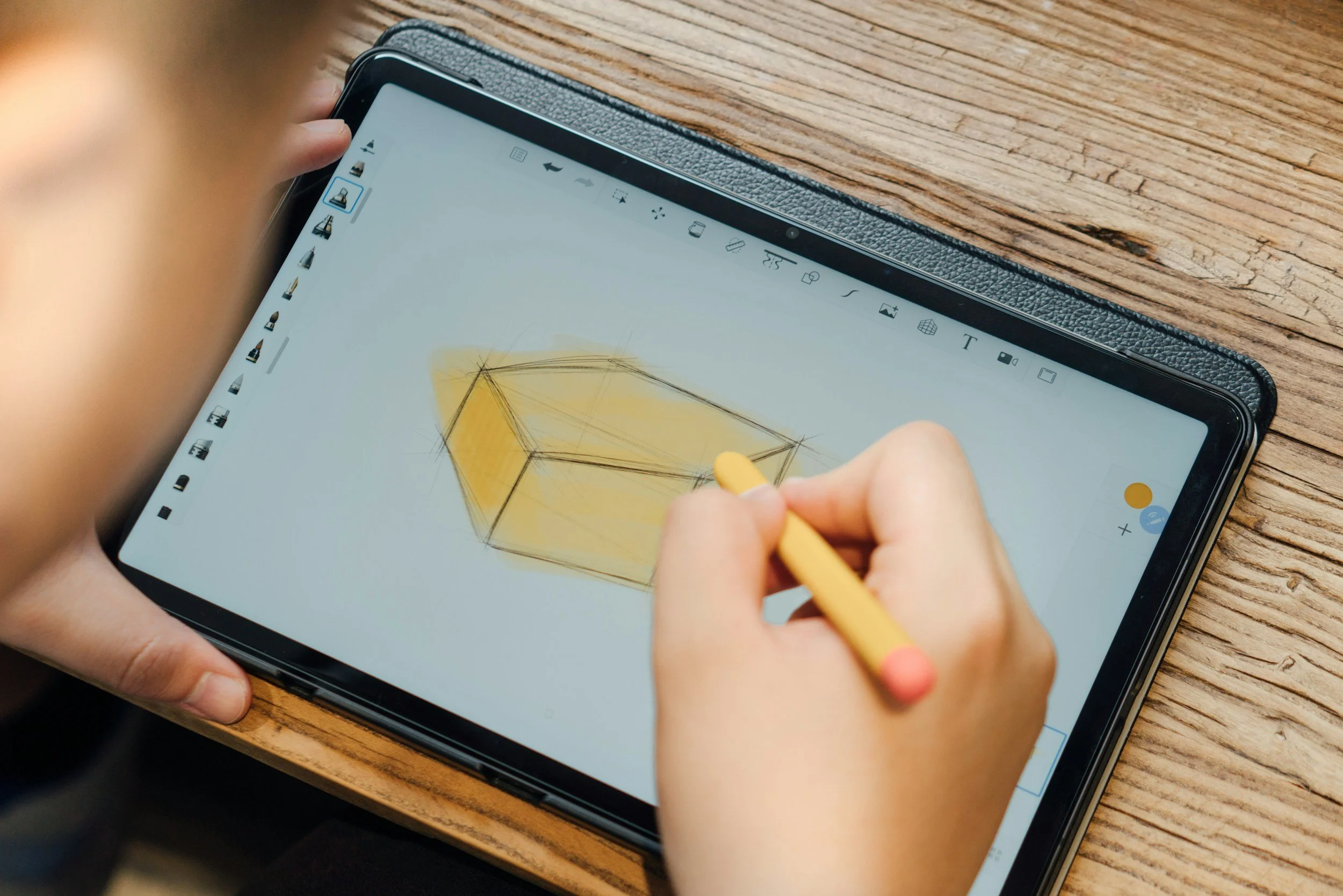 Person drawing a geometric cube on a tablet with a stylus, resting on a wooden surface.