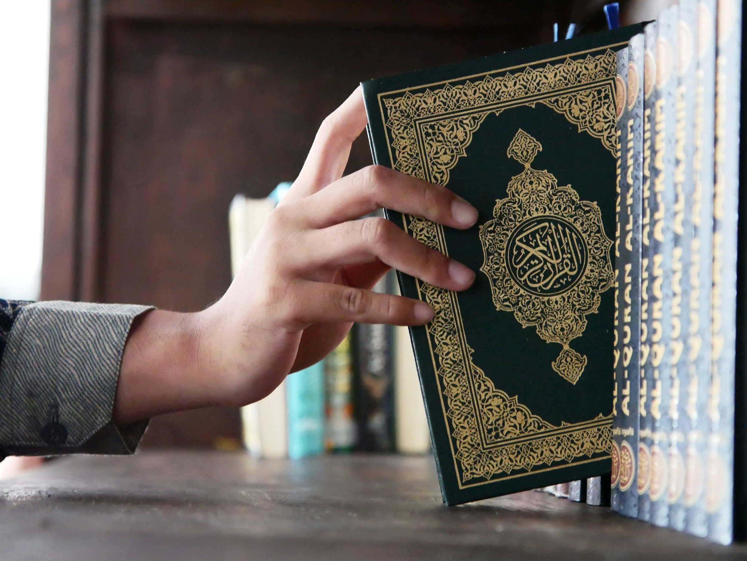 A person holding a green and gold decorated book, standing in front of a bookshelf with other books.