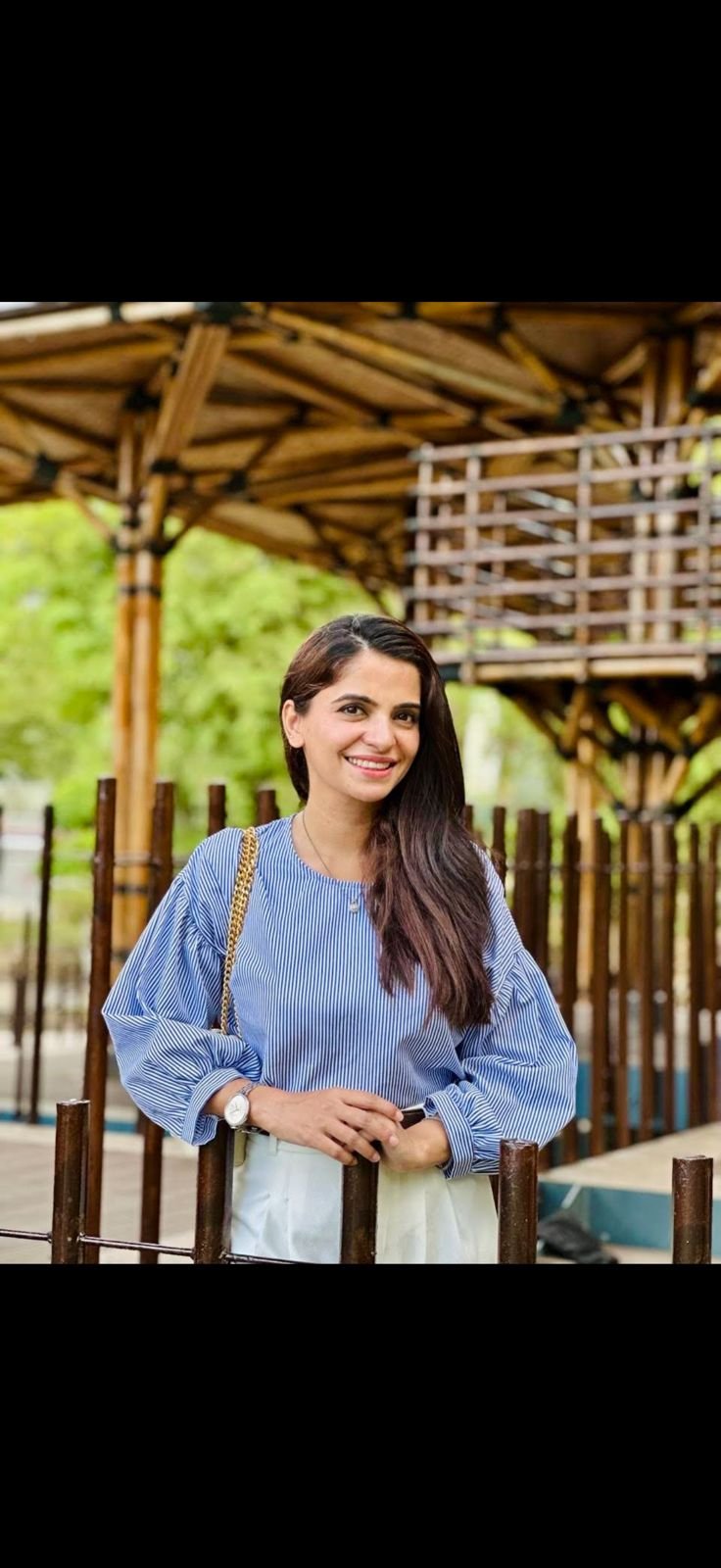 A woman with long dark hair, wearing a blue and white striped blouse, smiling and standing outdoors near a wooden fence with a bamboo structure and green trees in the background.