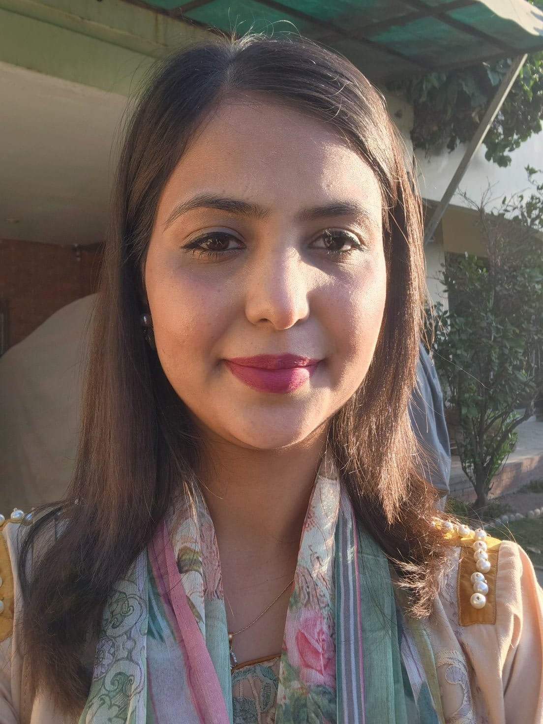 A woman with medium-length dark hair, wearing a colorful saree with pearl embellishments on the shoulder, standing outdoors during daylight with a building and trees in the background.