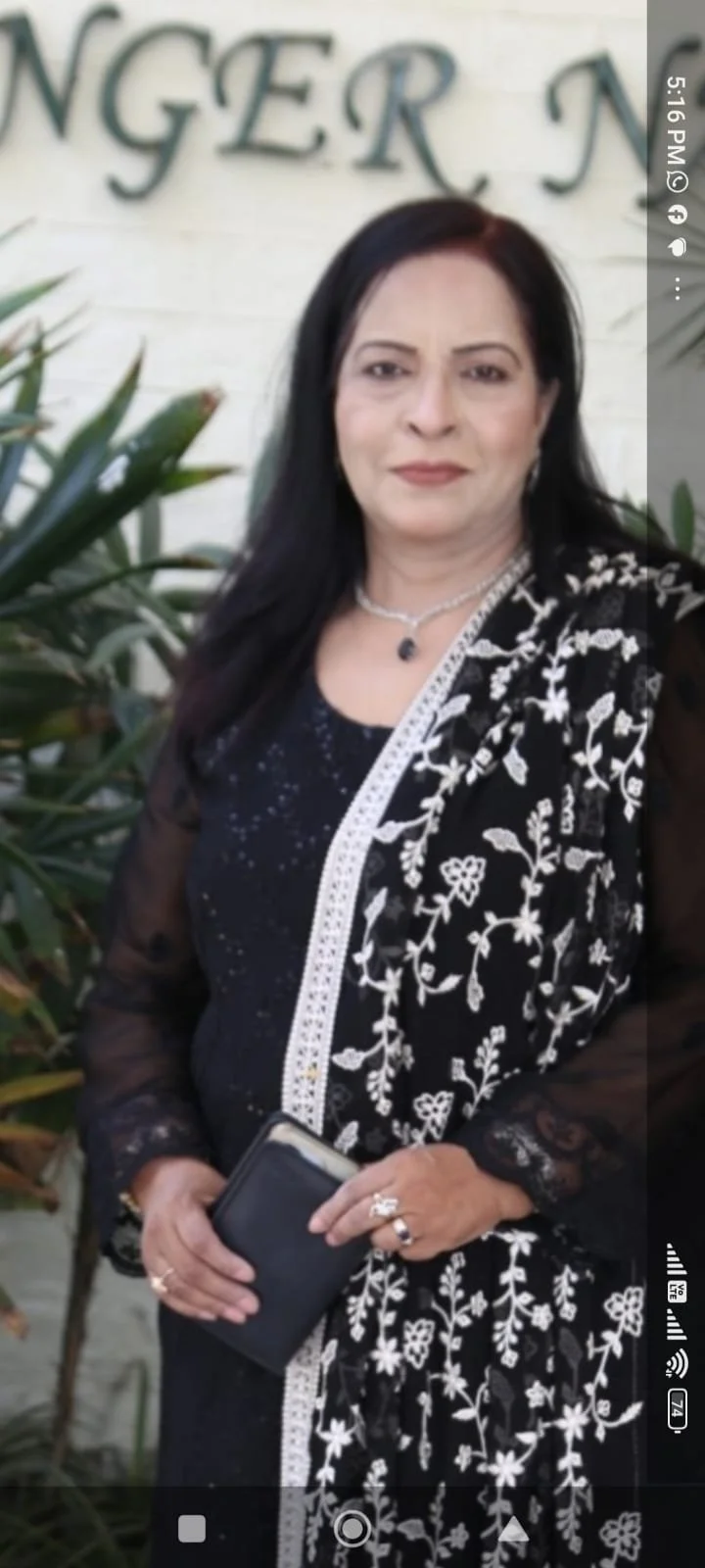 A woman with black hair wearing a black dress with white floral embroidery and a sheer black shawl holding a phone, standing in front of a white wall with green plants to the side.