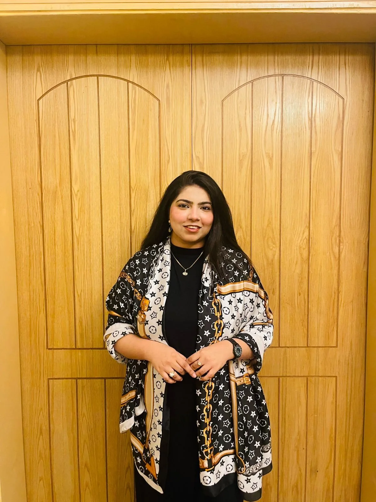 A woman with long dark hair stands in front of a wooden door, wearing a black top and a patterned shawl with black, white, and gold accents. She is smiling and has jewelry on, including a necklace, earrings, a watch, and rings.