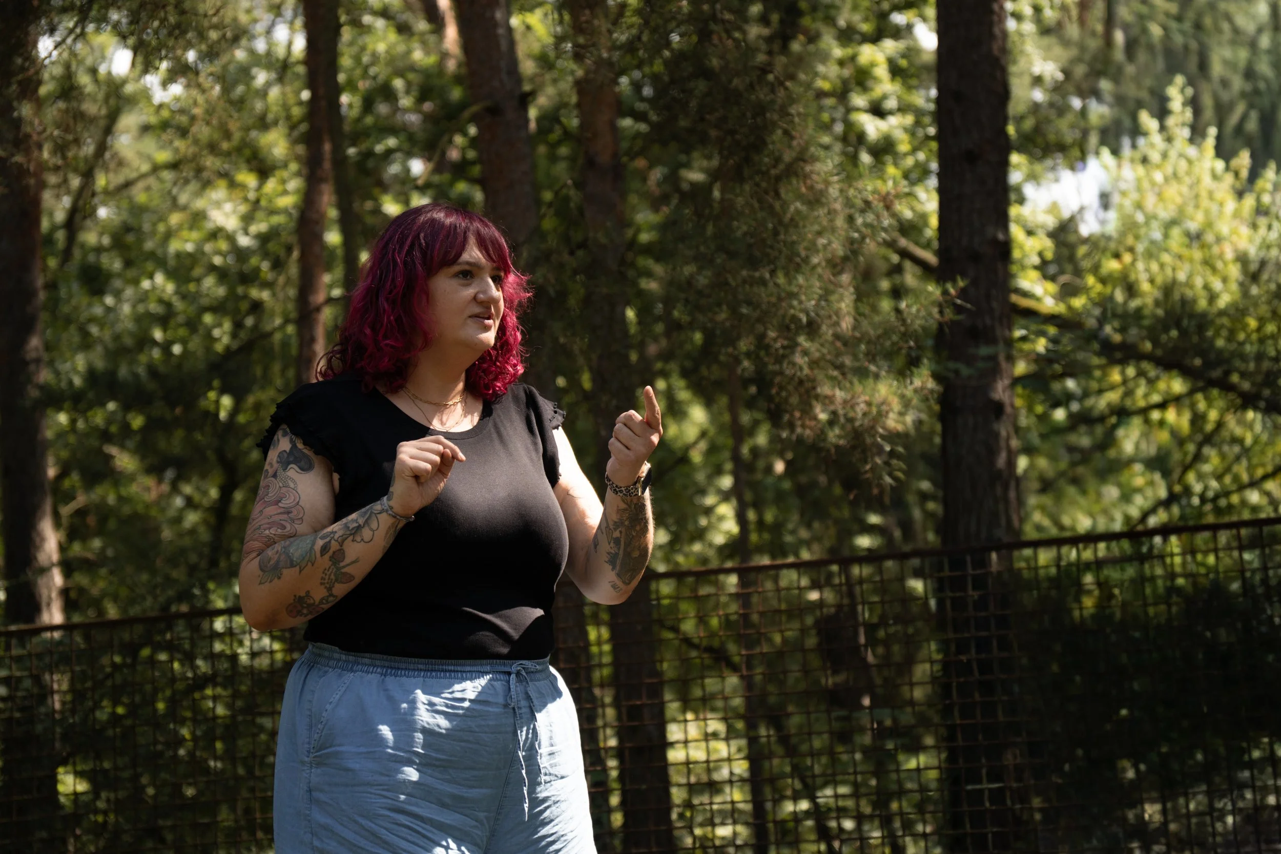 Eine Frau mit pink gefärbtem, lockigem Haar, schwarzen T-Shirt und Jeans-Hosen läuft im Park.