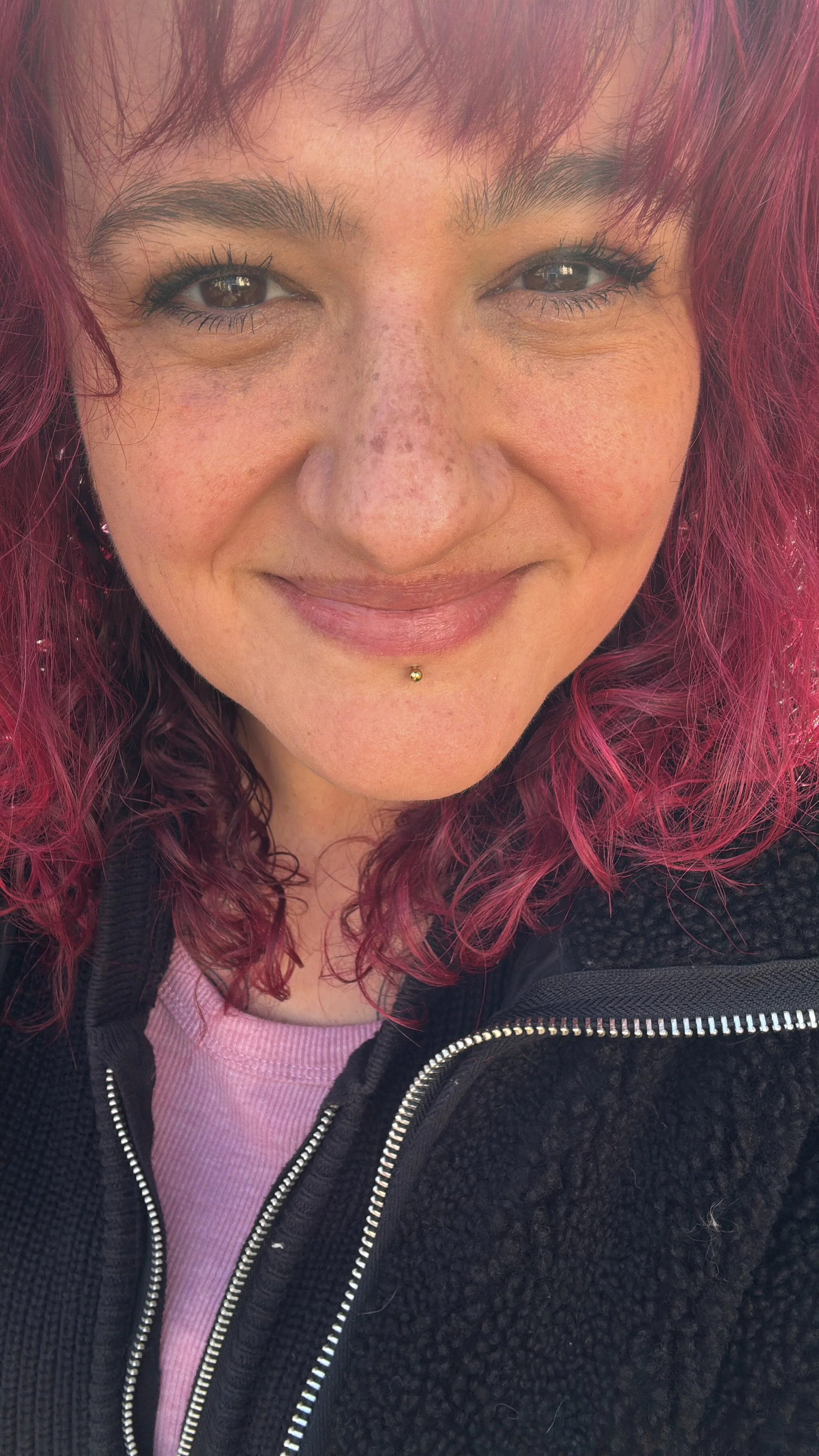 Nahaufnahme eines lachenden Gesichts einer Frau mit roten, lockigen Haaren, Sommersprossen und Piercing unter der Unterlippe, trägt einen schwarzen Pullover mit Reißverschluss und einen rosa Oberteil.