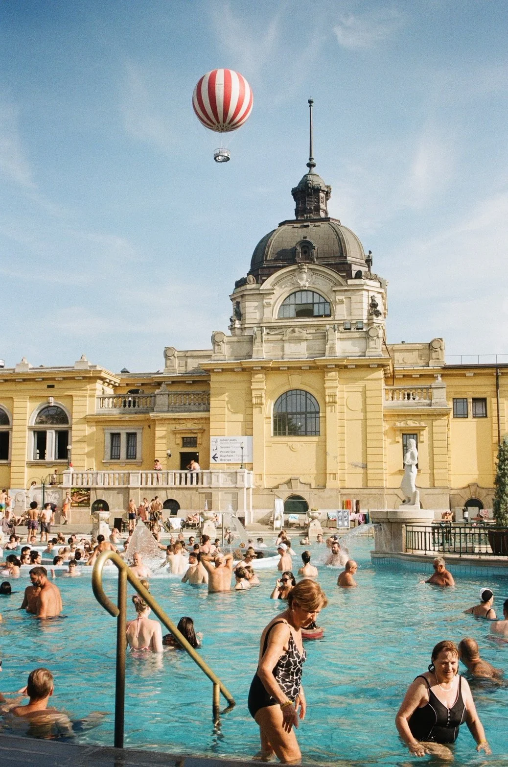 People swimming and relaxing in a large outdoor thermal bath with a historic yellow building and a hot air balloon in the sky.
