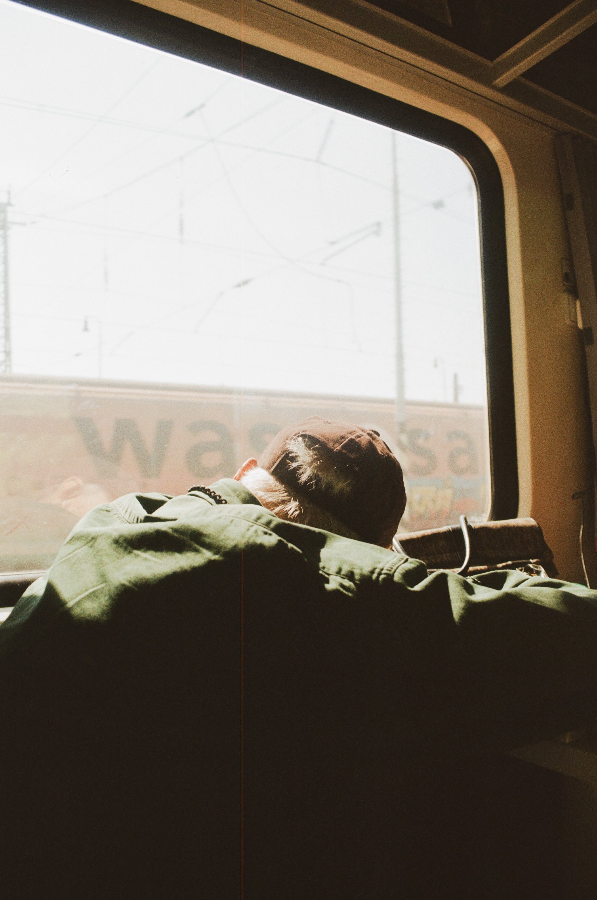 Person lying on a train seat, wearing a brown cap and a green jacket, with head resting on the window side, and the outside view of railway power lines and sky visible through the window.