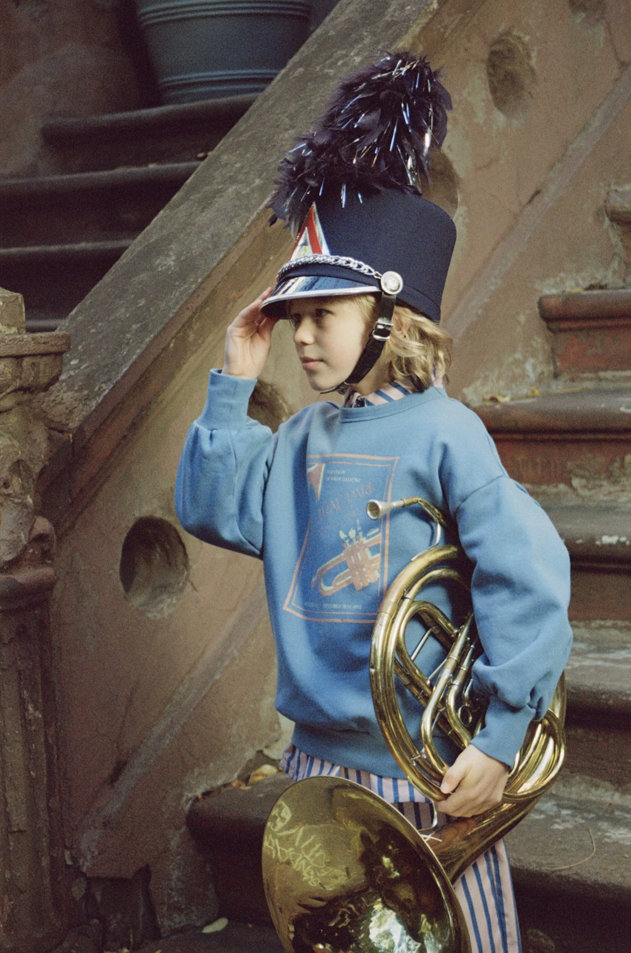 Young boy wearing a hat with feathers, holding a brass musical instrument, standing against a brick and stone wall.