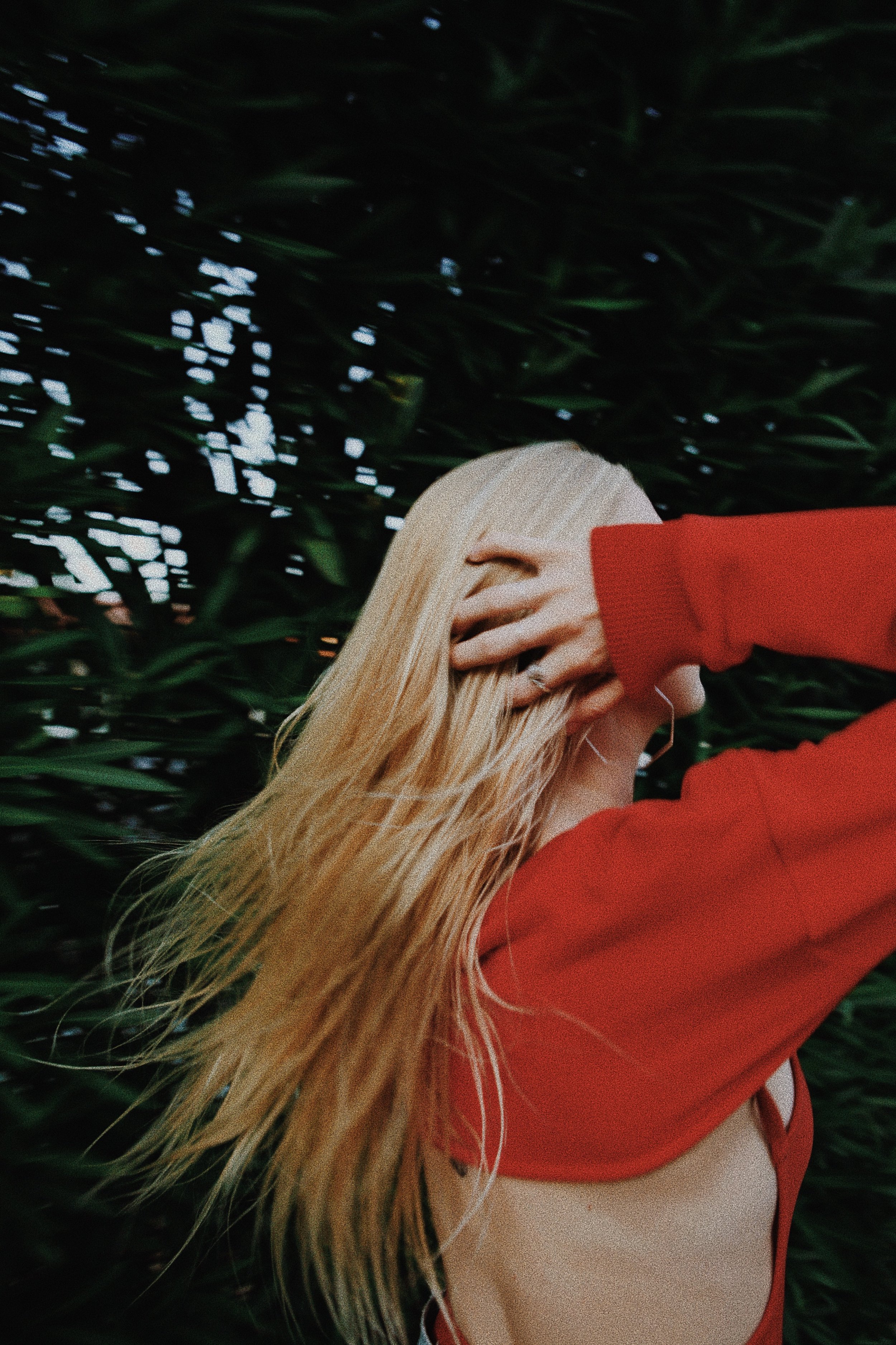 A woman with long blonde hair wearing a red jacket standing in front of greenery.