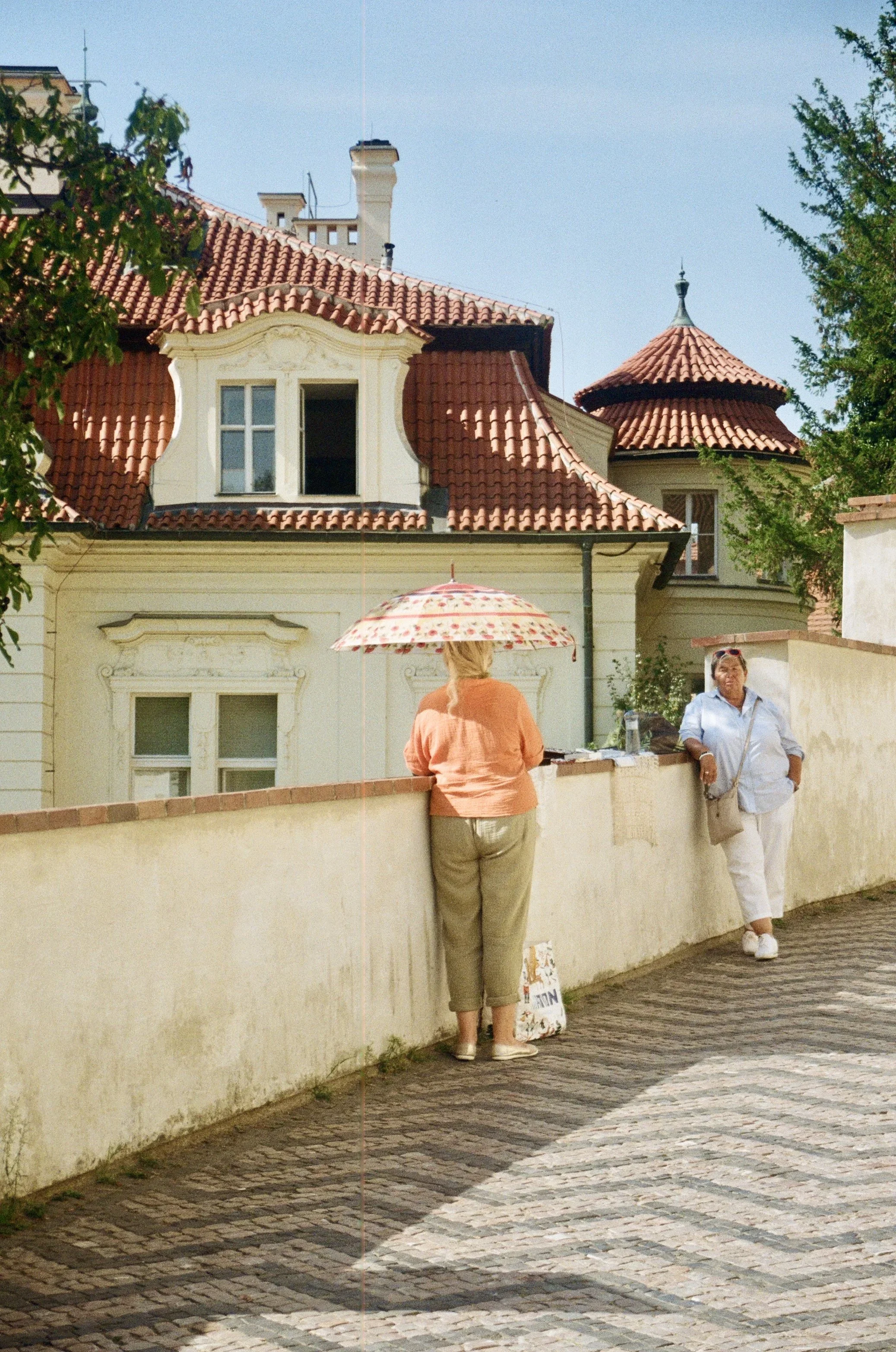 Two women standing by a brick wall in front of a yellow building with red-tiled roof and ornate windows, one woman with a floral umbrella and the other smiling in white shoes and pants.