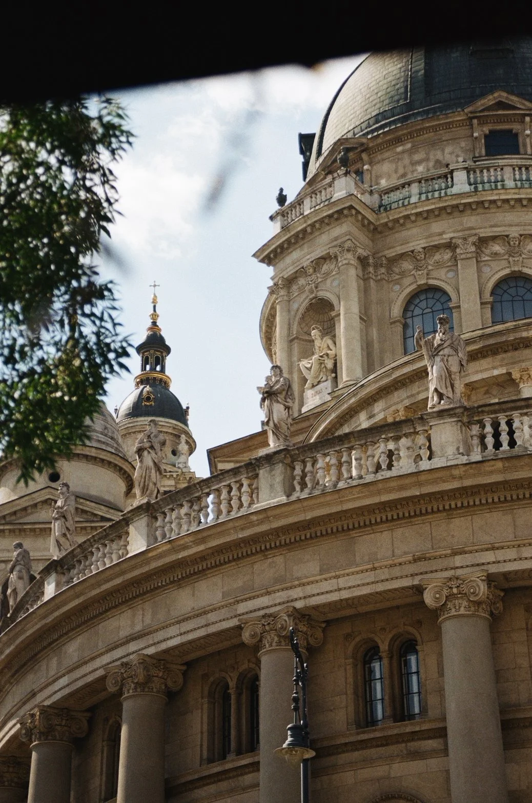 Close-up of a historic European cathedral's domed roof, ornate statues along the balustrade, and architectural details with a cloudy sky in the background.