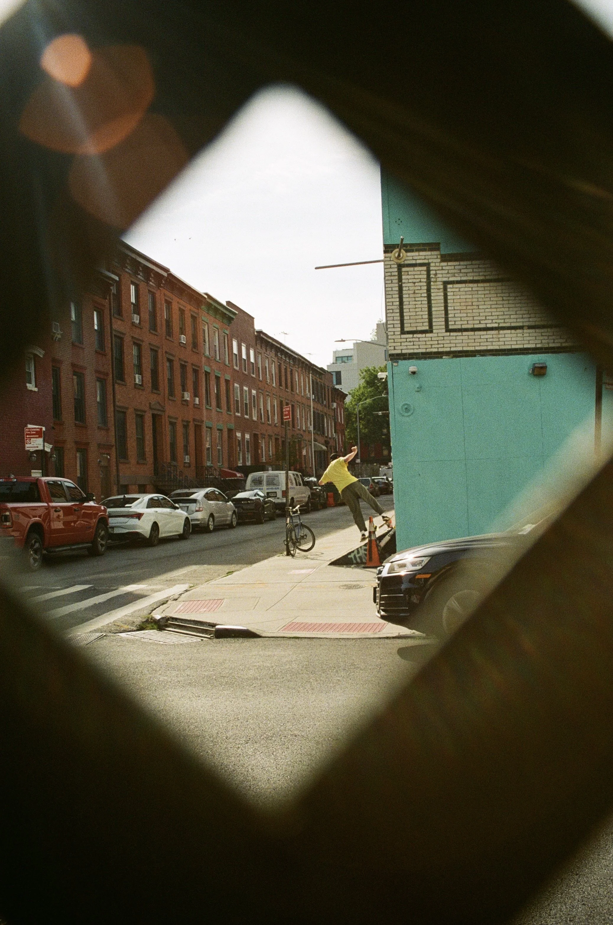 Street scene viewed through a small, diamond-shaped opening, showing a man riding a bicycle on the sidewalk with parked cars and brick buildings in the background.