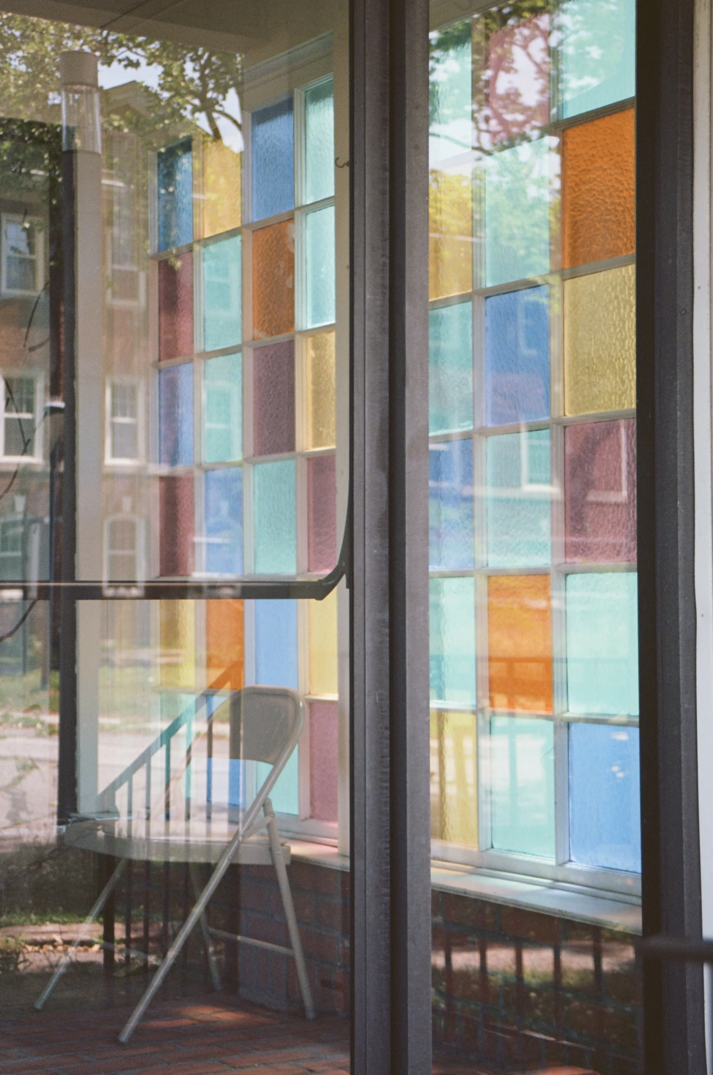 View through a window with colored stained glass panels and a white folding chair outside on a brick porch.